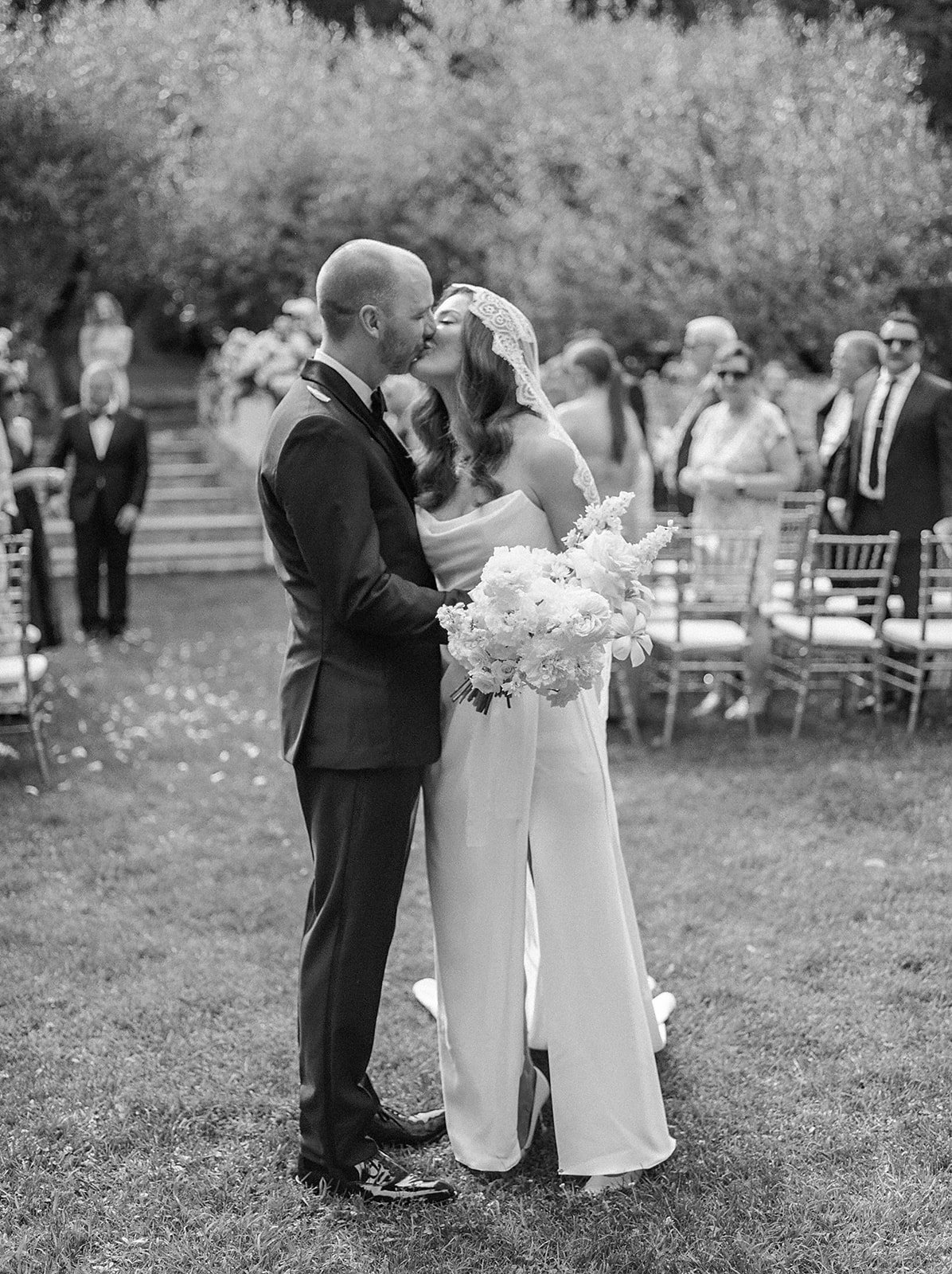 Long outdoor dining table decorated with white flowers and set with plates and glasses, surrounded by gold chairs, under string lights, in a garden setting.