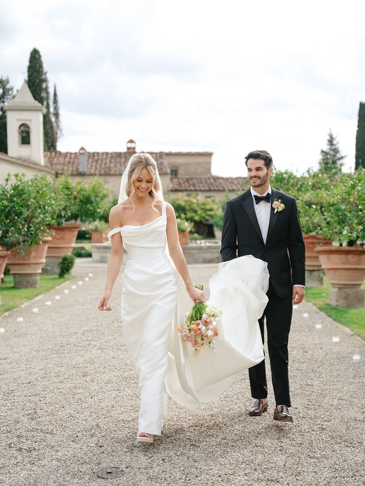 A bride in a white wedding gown and veil is seated on a vintage chair, smiling, with a groom in a black tuxedo standing behind her against a dark, artistic background.
