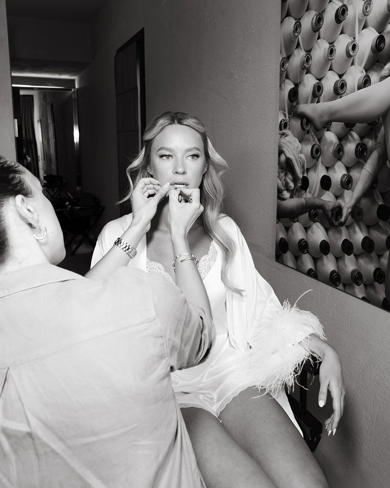 A makeup artist applies lipstick to a young woman sitting in a salon or dressing room, with rolls of fabric or material stacked on the wall behind them.