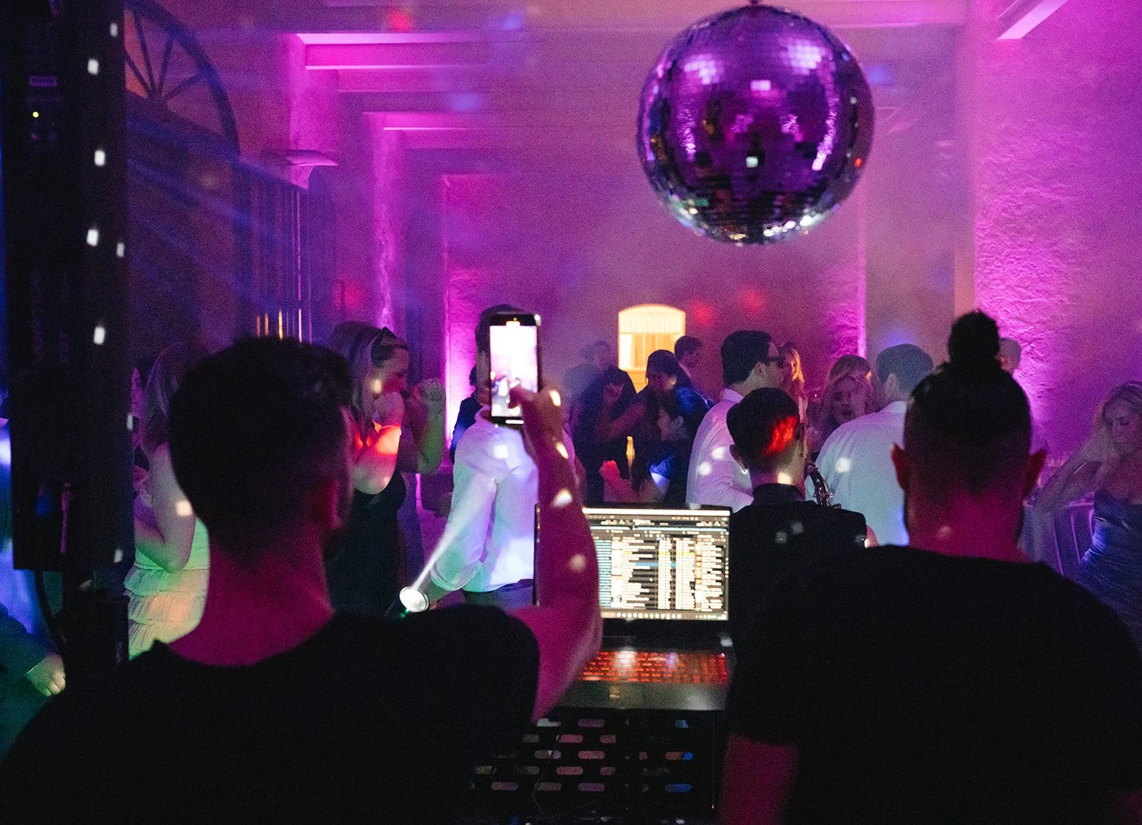 People dancing at a nightclub with purple lighting, a disco ball hanging from the ceiling, and a DJ playing music. Some individuals are taking photos, and the scene is lively and colorful.