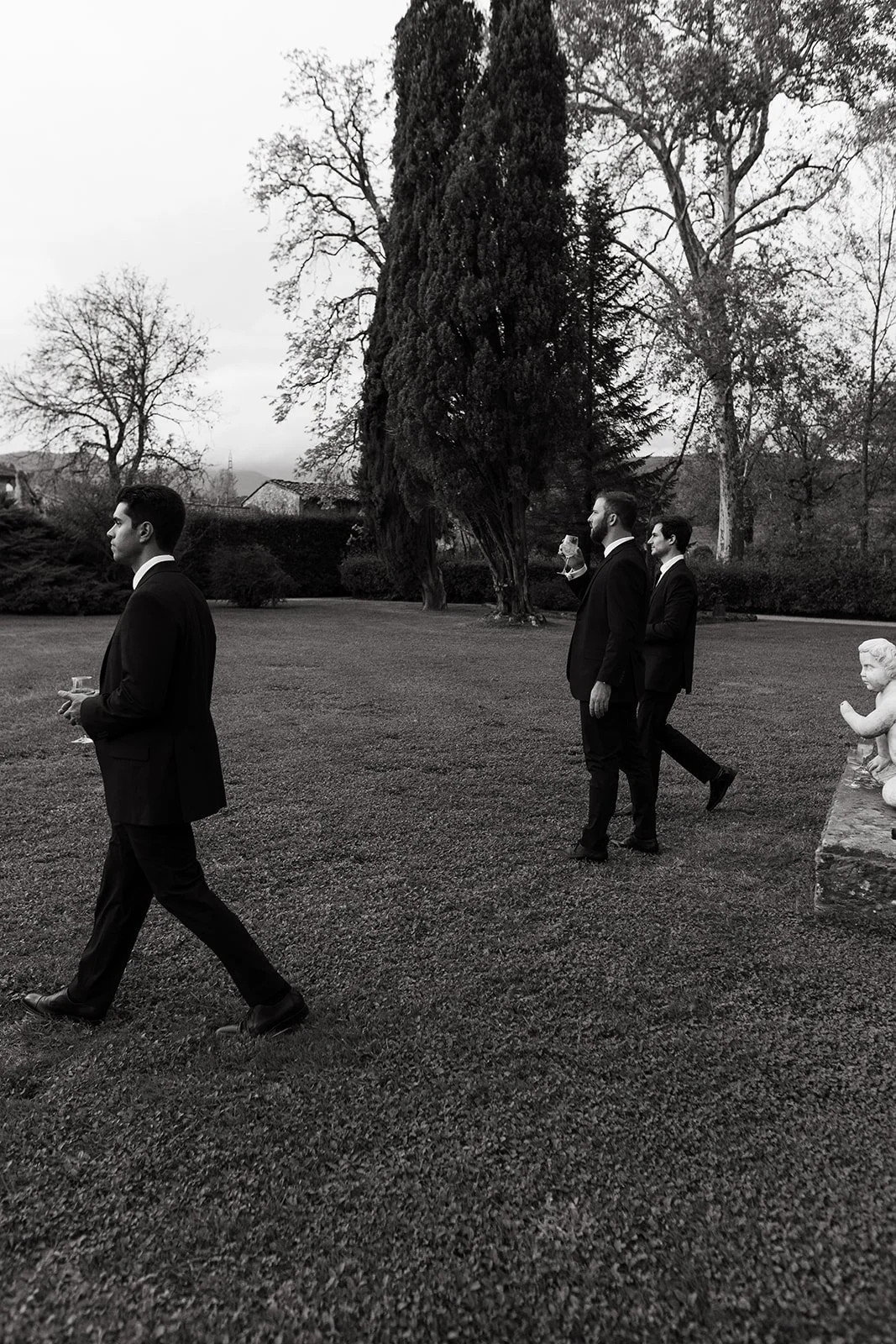 Four men dressed in black suits walking outdoors in a park with trees and a stone sculpture of a child, one man holding a drink, the scene is black and white.