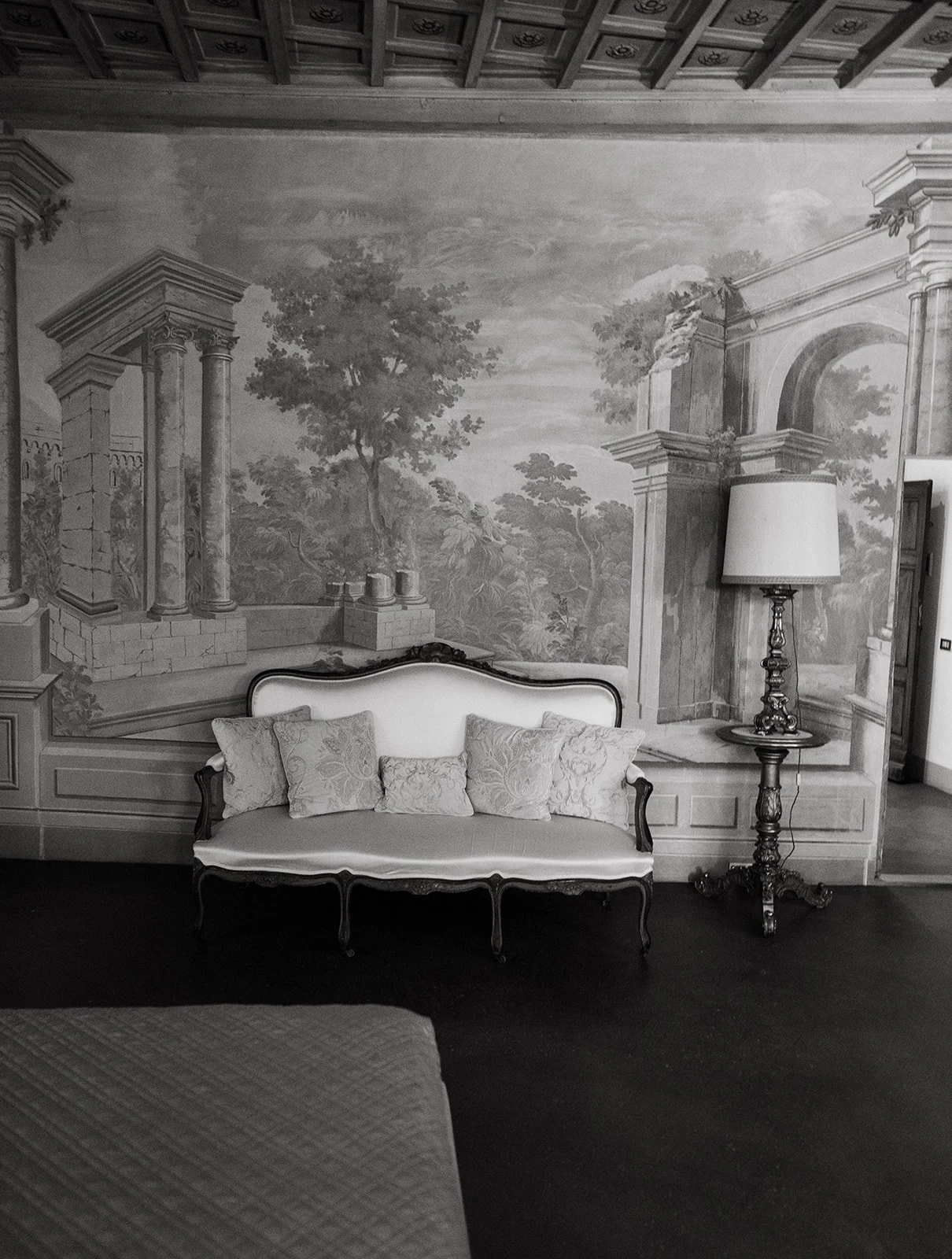 A black and white photo of a vintage style sofa with four decorative pillows, against a large mural wall depicting classical architecture and trees, with a floor lamp beside the sofa.