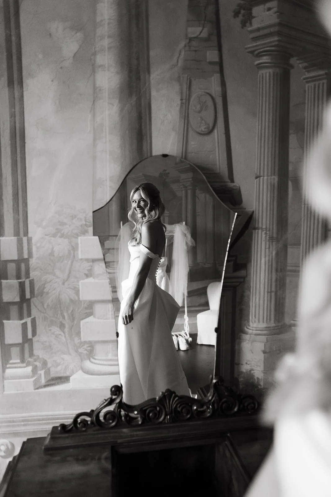 A woman in a wedding dress is looking at her reflection in an ornate mirror, smiling.
