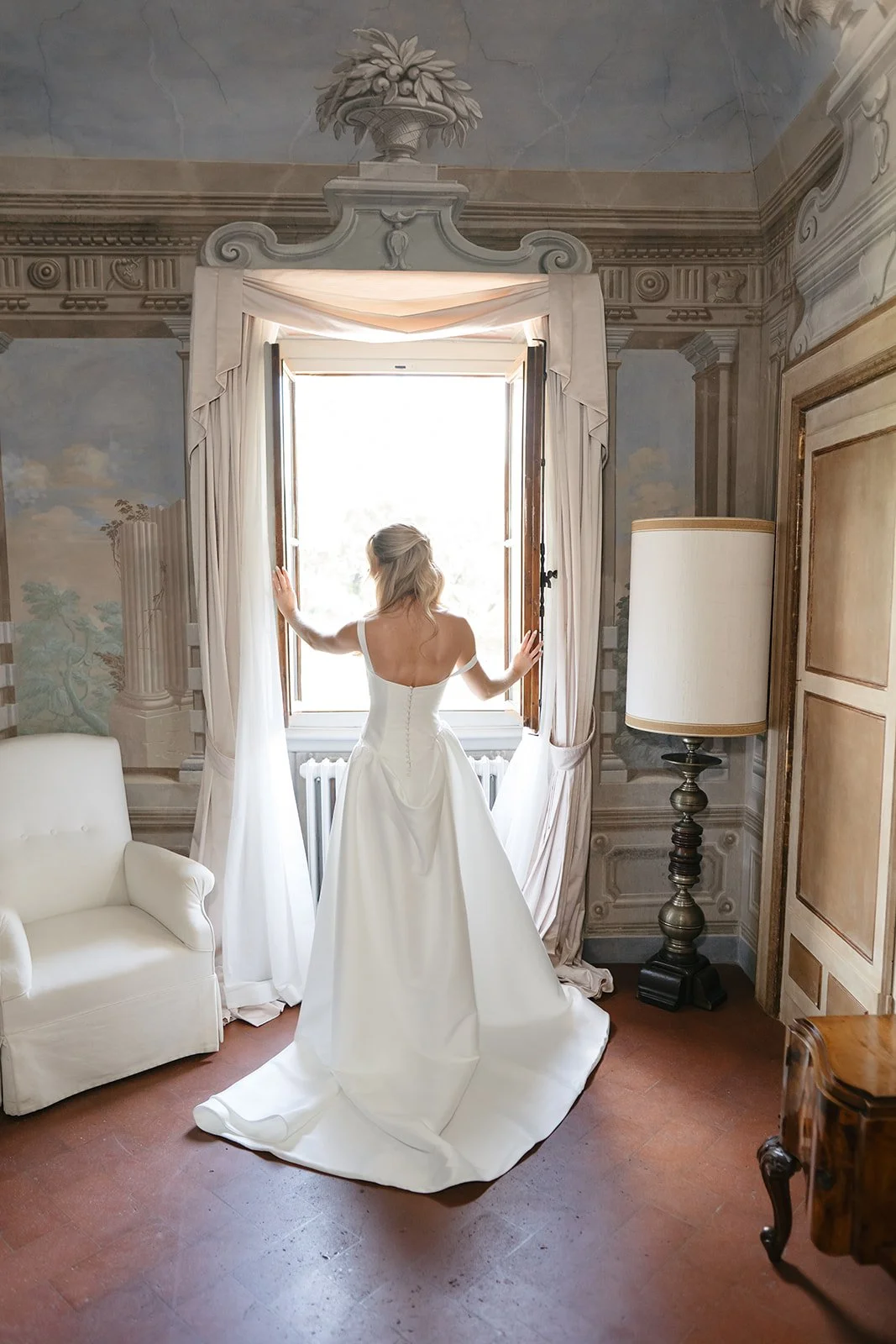 A bride in a white wedding dress stands by an open window, looking outside, with sunlight shining in, in a room with ornate wall decorations, a white armchair on the left, and a large lamp on the right.