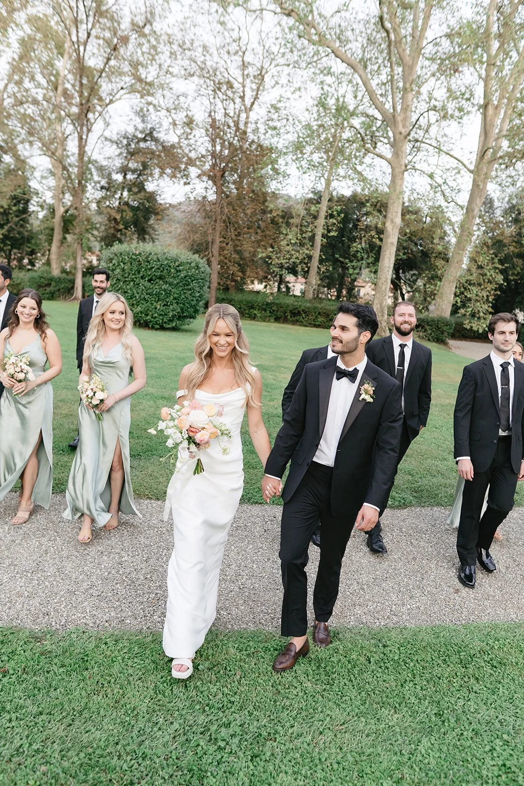A wedding party walking outdoors on a grassy area with trees in the background. The bride and groom are leading, holding hands, with the bride in a white gown and the groom in a black tuxedo. Bridesmaids in light green dresses and groomsmen in black 