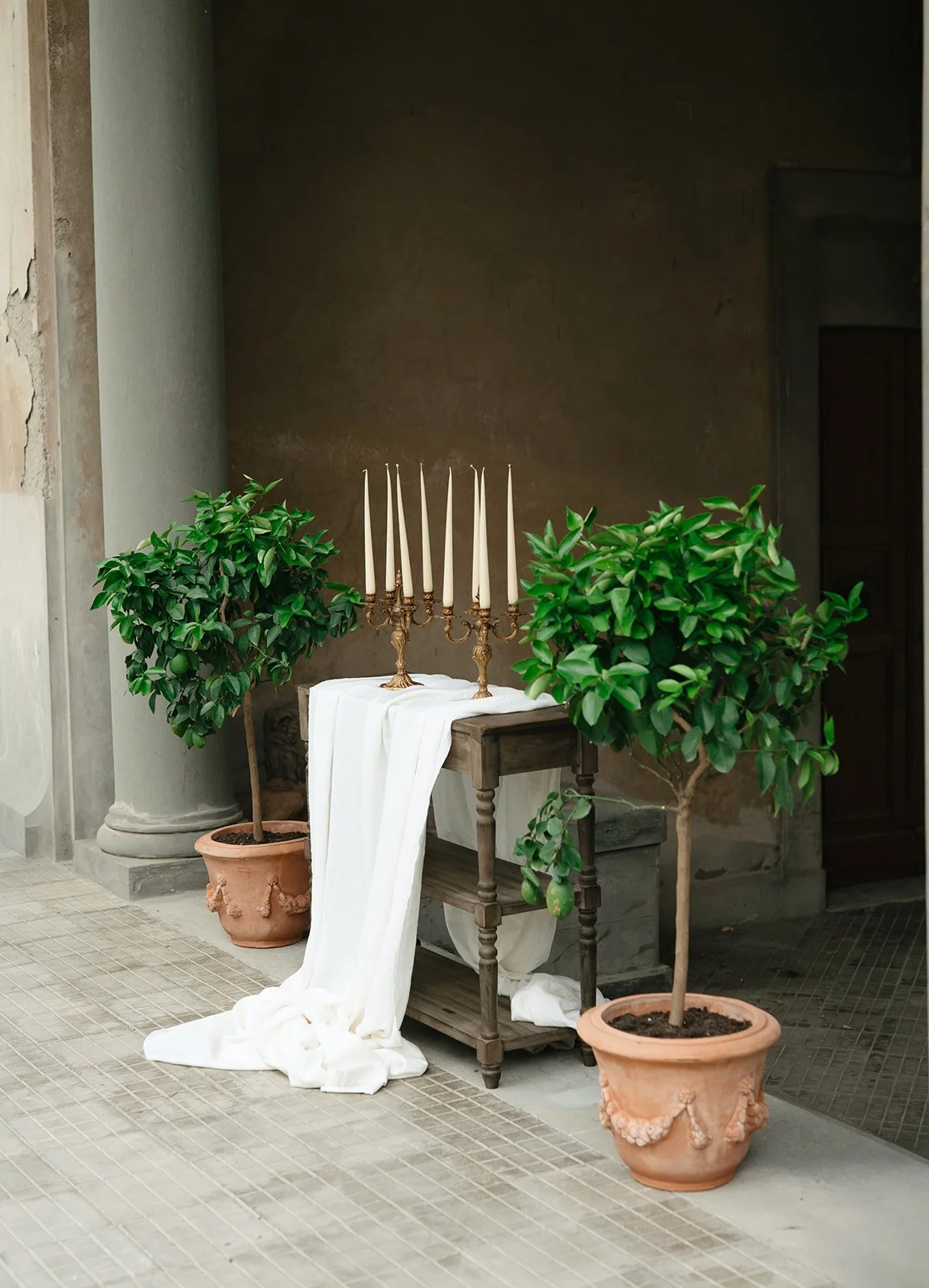An outdoor scene with two potted trees, a small wooden table holding a candelabra with nine unlit candles, and white fabric draped over the table and extending to the ground. The background includes a wall and doorway.