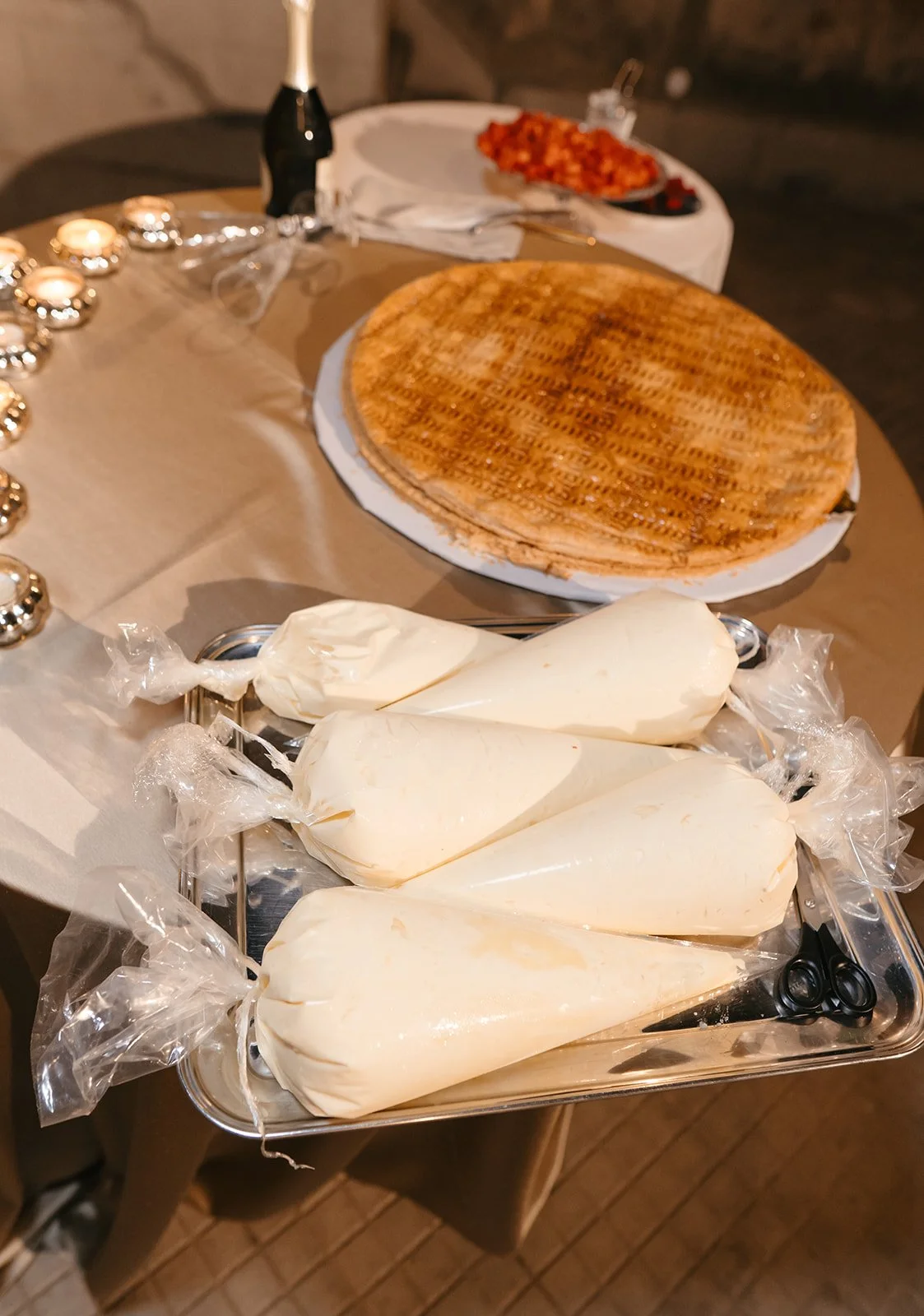 A table set for a celebration with a round cake, a tray of four pastry bags filled with white frosting, a bottle of champagne, and a platter of sliced meat or ham. There are tealight candles and napkins with cutlery.