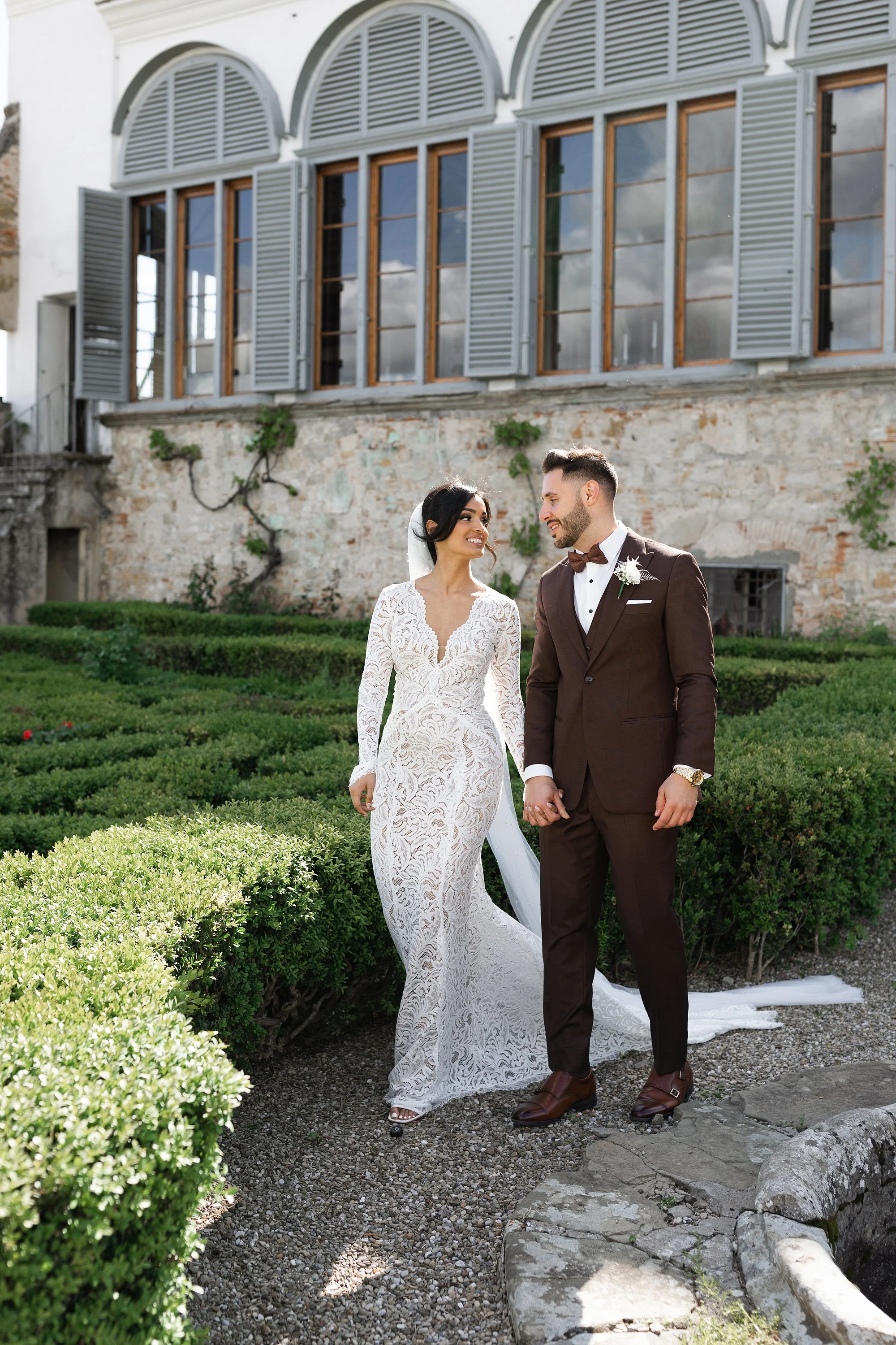 A couple in wedding attire holding hands, walking outdoors under a floral arch, with greenery in the background, during daytime.