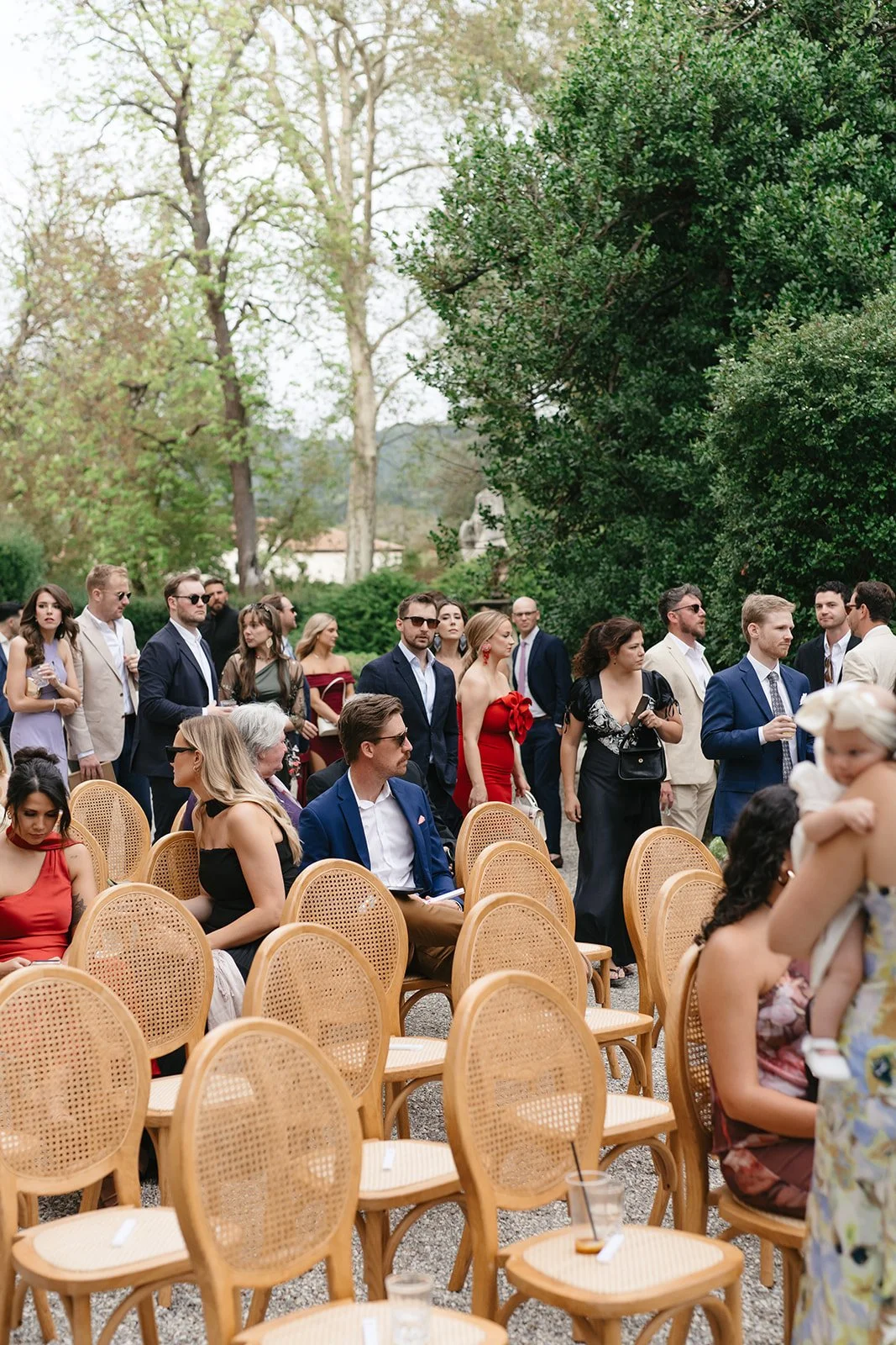 People attending an outdoor event, with some seated on wooden chairs and others standing, in a lush green garden setting.