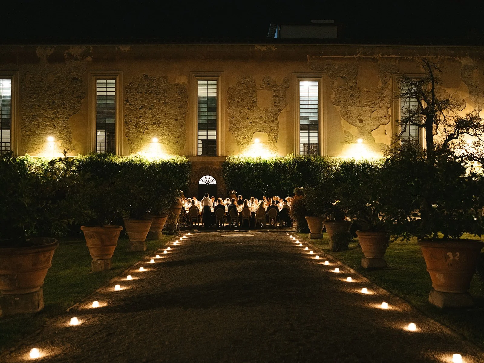 Nighttime scene of an outdoor dinner event with a long candlelit pathway leading to a gathering of people seated at a long table against an old stone building, illuminated by warm lights.
