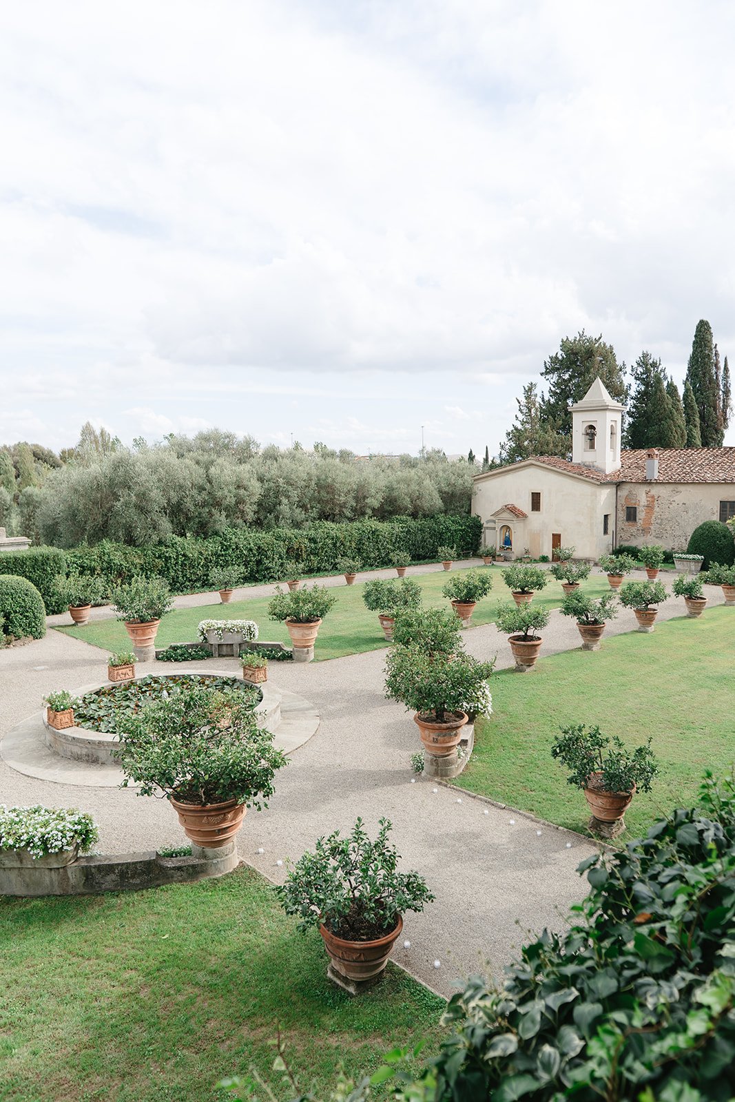 A scenic view of an Italian garden with potted plants and greenery, a small church building in the background, under a partly cloudy sky.