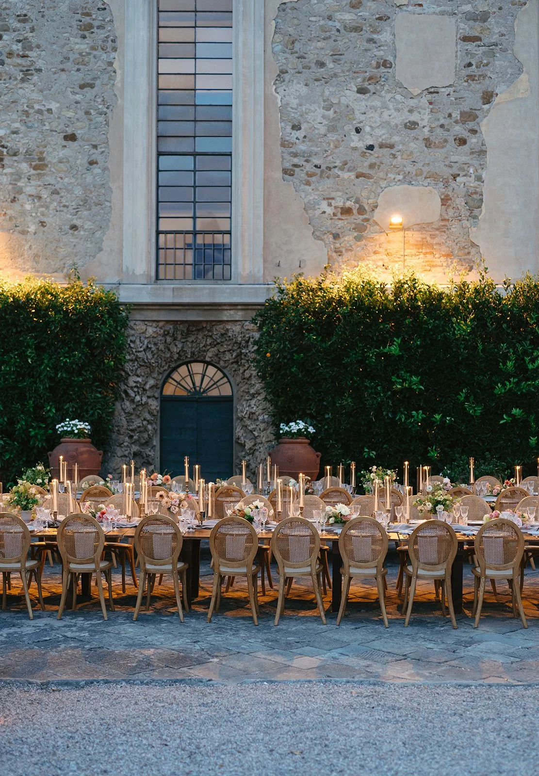Outdoor dining table set up with floral centerpieces and candles, located in a courtyard with stone walls, greenery, and a tall window.