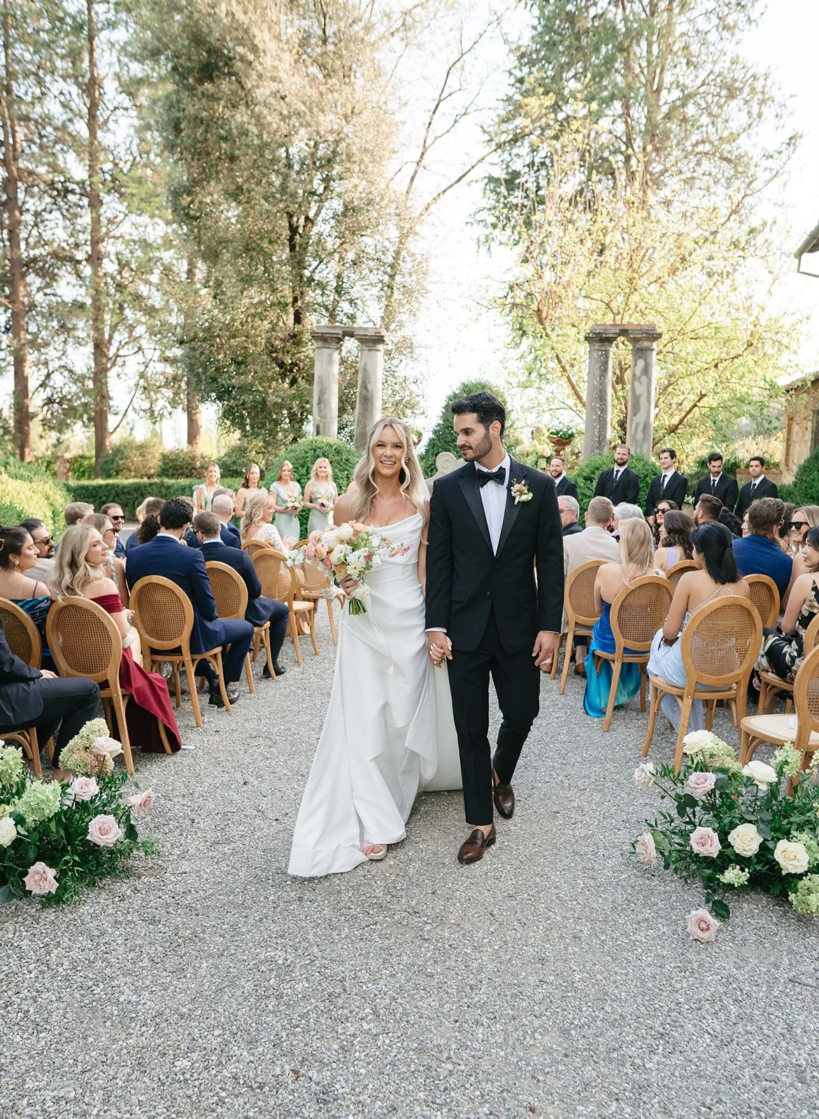 A bride and groom walking hand in hand at their outdoor wedding ceremony, surrounded by guests on wooden chairs, with bridesmaids and groomsmen in the background and green trees.