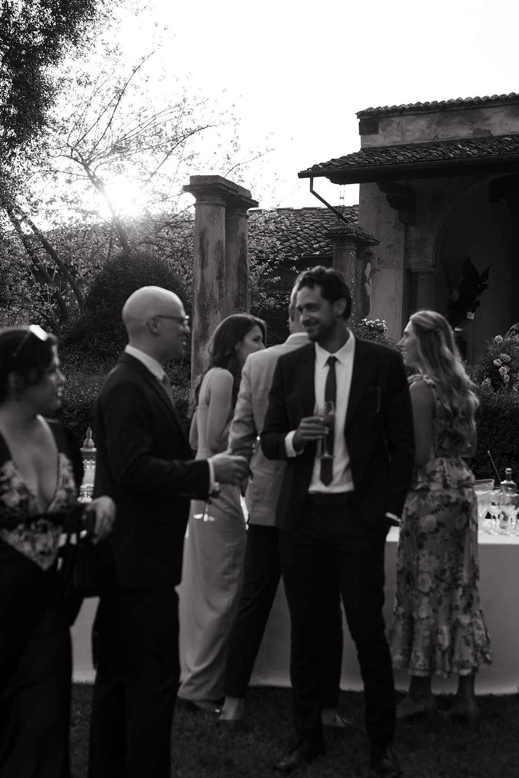 People gathered outdoors at an evening event, some holding drinks, with a historic building and trees in the background.