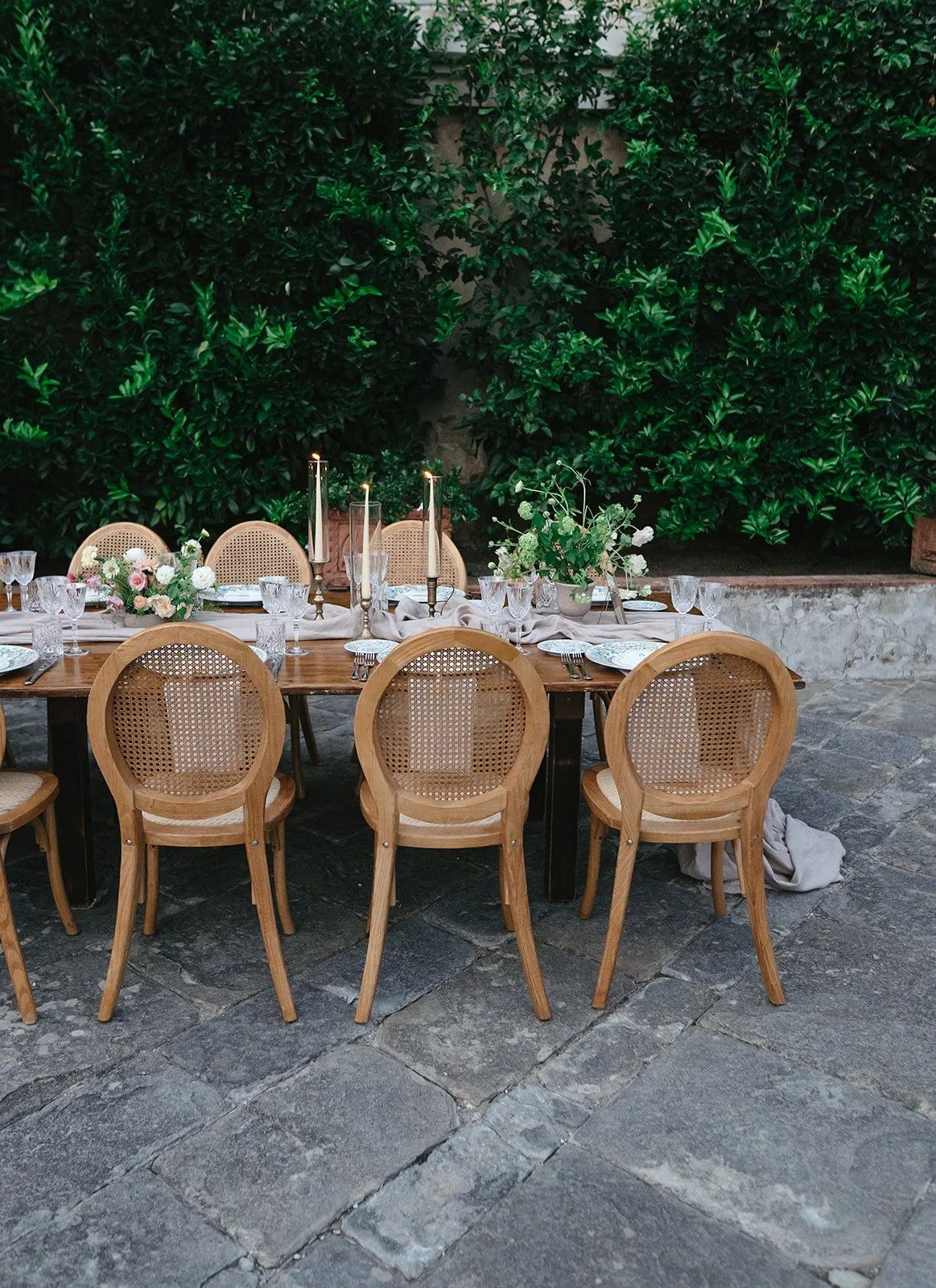 An outdoor dining table set with glassware, plates, cutlery, and floral centerpieces, surrounded by wooden chairs, against a backdrop of green shrubbery and stone pavement.