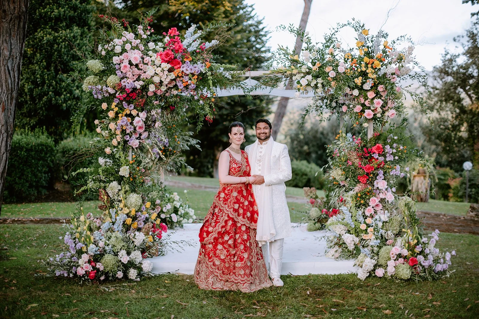 A couple dressed in traditional Indian wedding attire stands under a large floral arch in a garden. The woman wears a red and gold embroidered lehenga, and the man wears a white sherwani with intricate patterns, both smiling and holding hands.