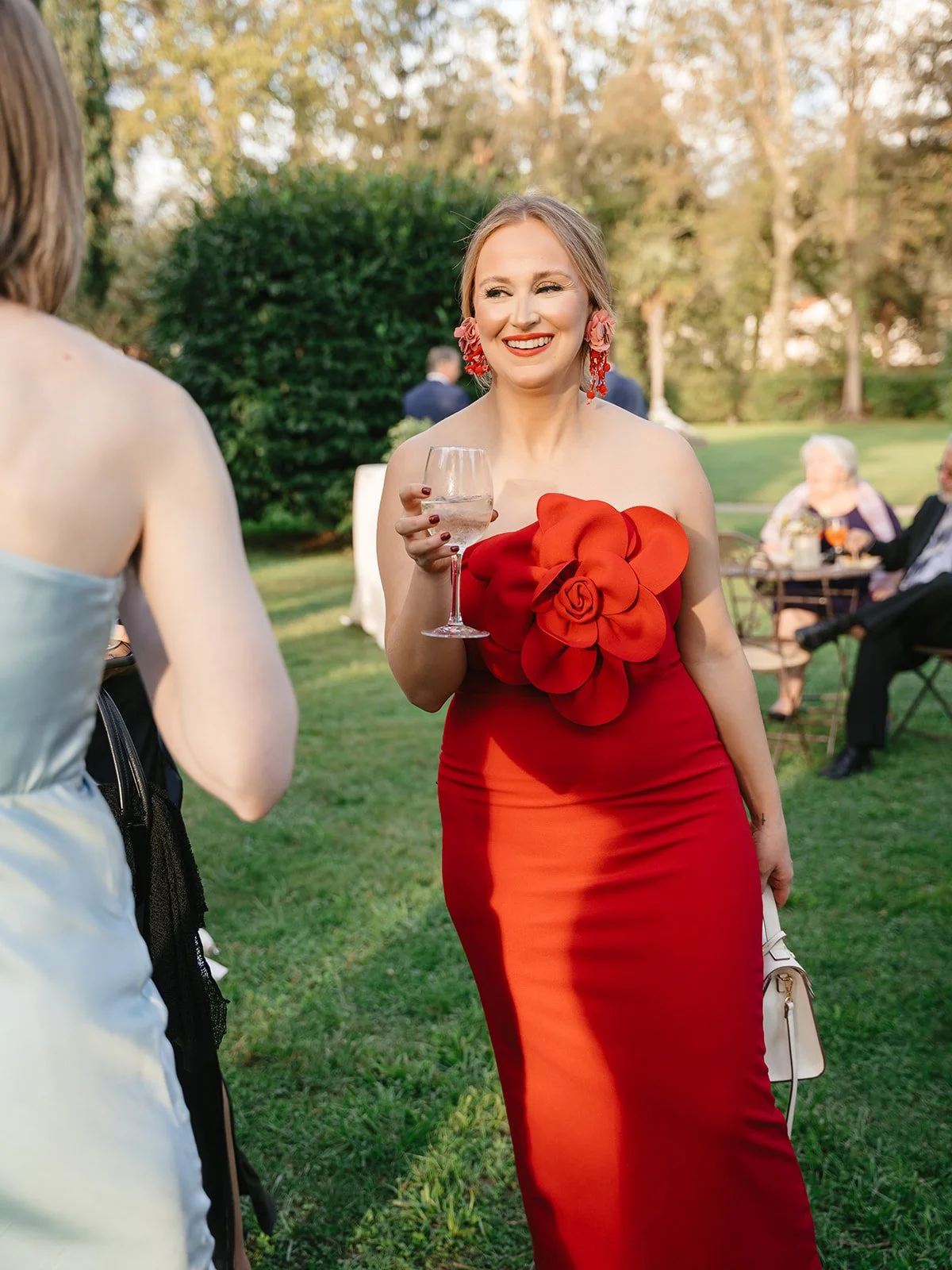 Woman in a strapless red dress with floral embellishments, holding a wine glass, smiling outdoors at a social gathering.