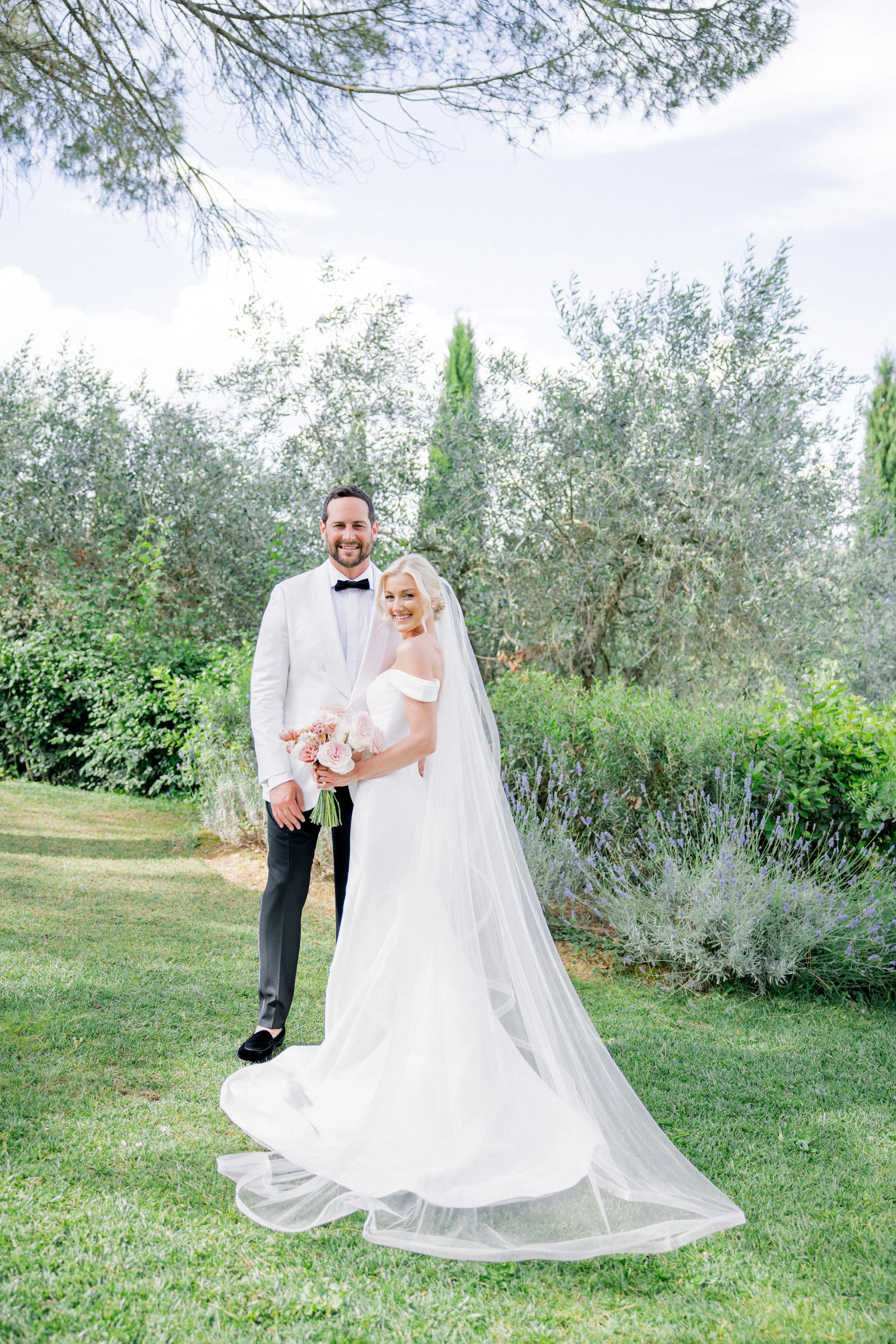 A flat lay of wedding invitation details, a watercolor house illustration, a Boston dog illustration with rings, floral decorations, and pink and white flowers on a light background.