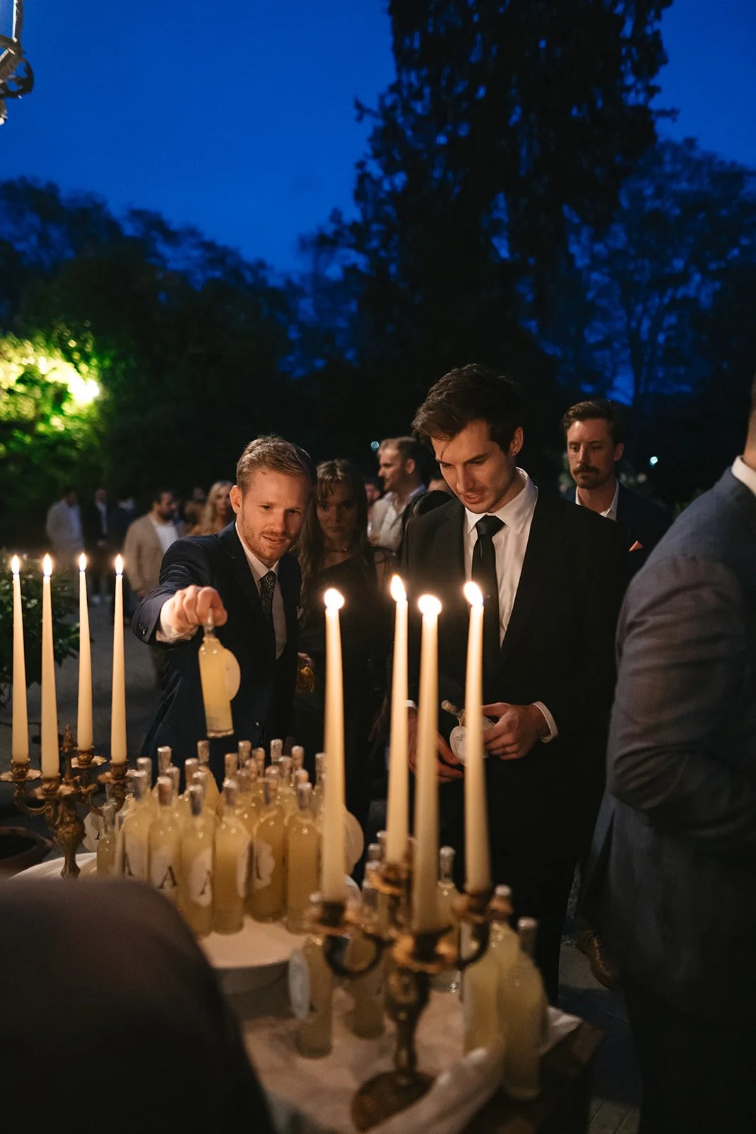 People in formal attire gather around a table with lit candles and bottles at night, outdoors, with trees in the background.