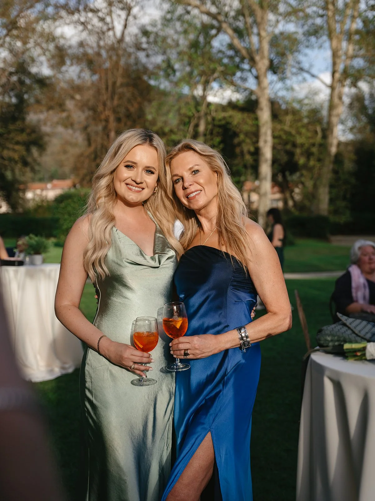 Two women in evening gowns holding glasses of orange-colored drink, standing outdoors at evening event with trees and tables in the background.