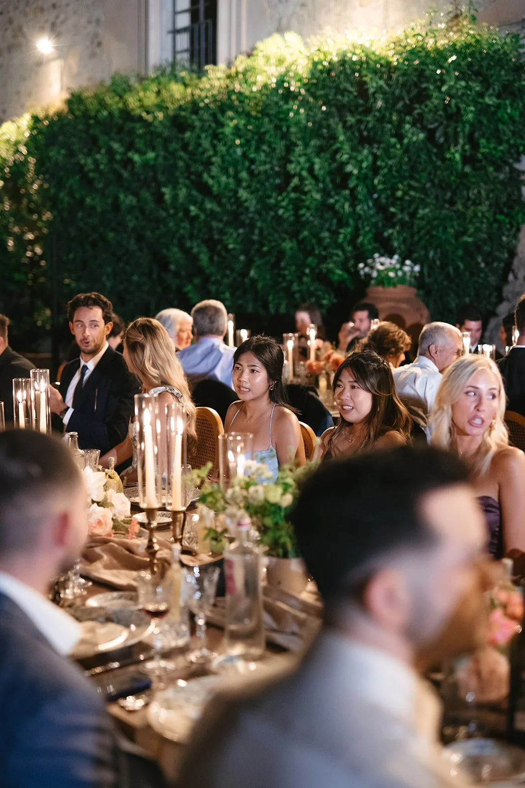 Guests seated at a formal dinner table outdoors with floral centerpieces and tall candles, under a large tree with green foliage, in an elegant evening setting.