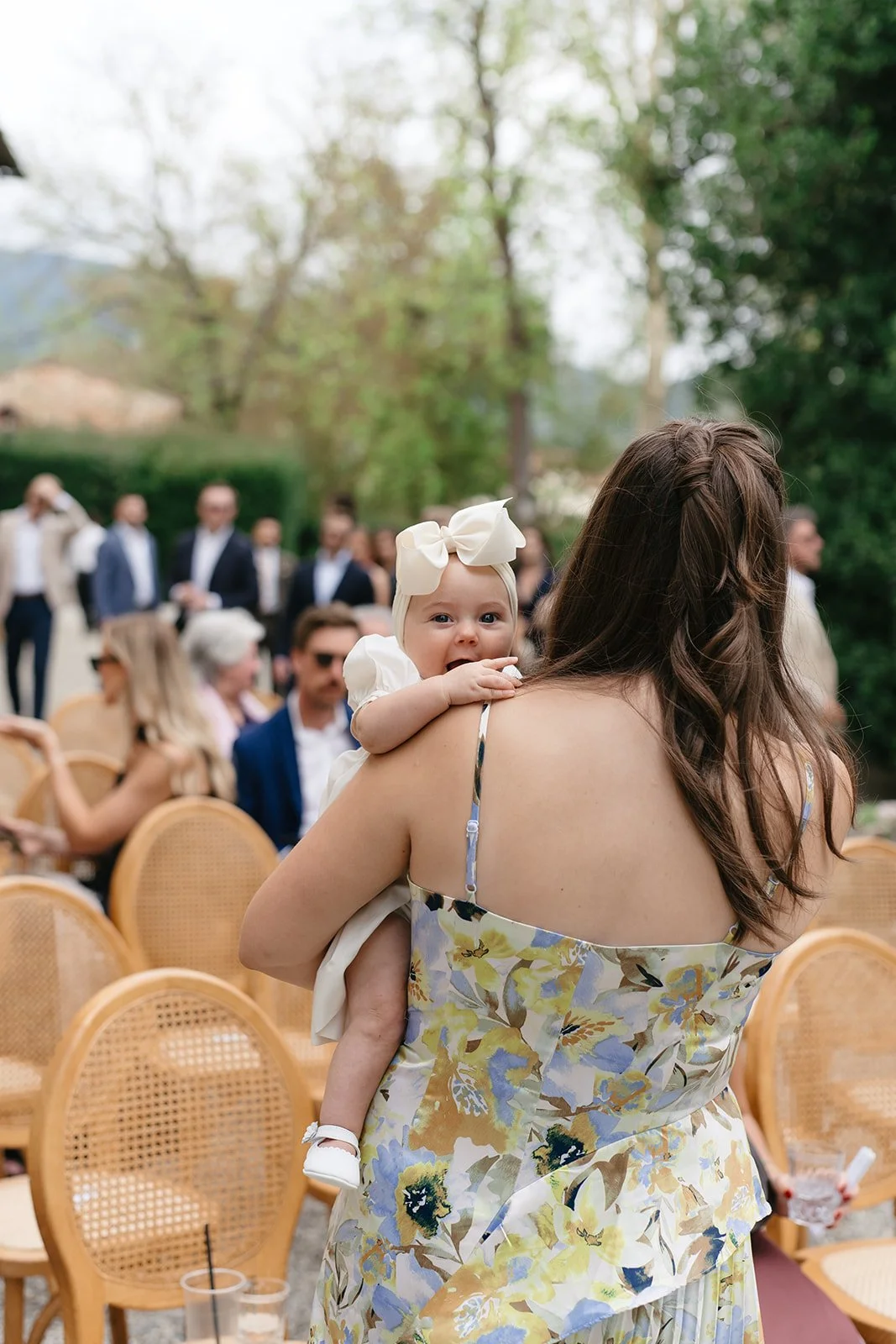 A woman holding a baby girl at an outdoor gathering, with several people and chairs in the background.