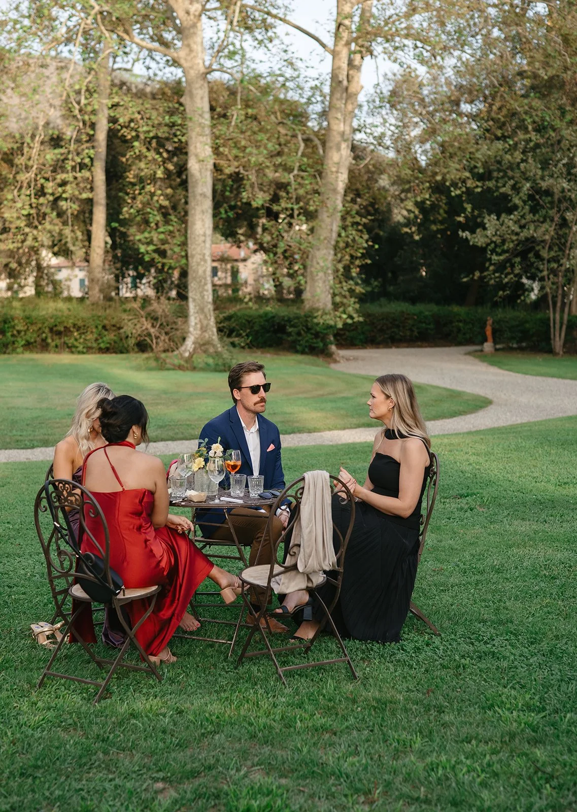 Four people are sitting around a table outdoors on a green lawn, with trees in the background. They are dressed formally, with one woman in a red dress, another woman in a black dress, and a man in a blue suit, some wearing sunglasses.