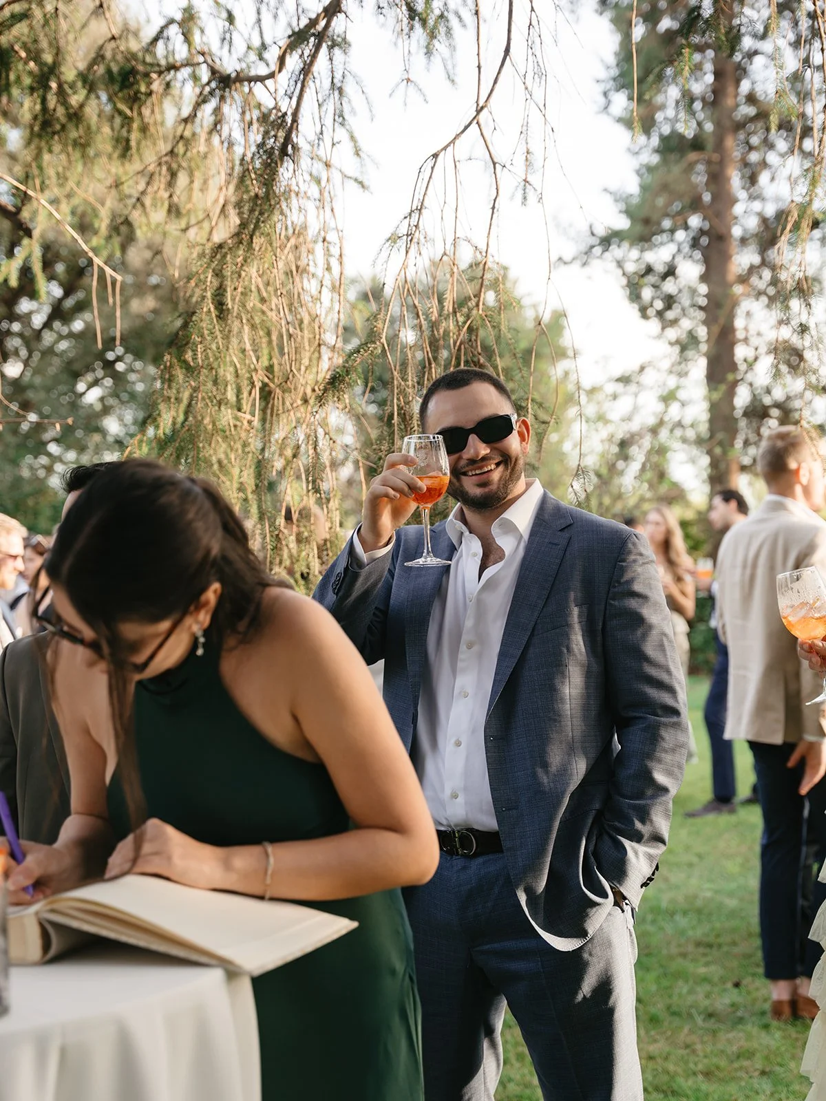 Man in a suit and sunglasses smiling and holding a glass of orange-colored drink at an outdoor gathering, with other guests in the background.