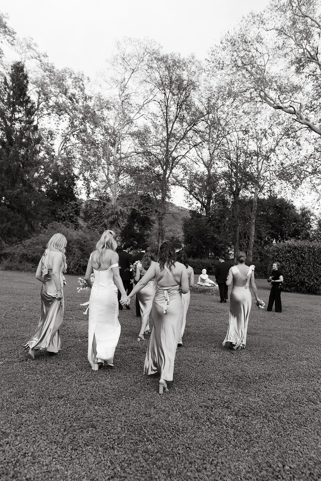 A group of women in long dresses walking outdoors in a garden setting, holding hands. The photo is in black and white.