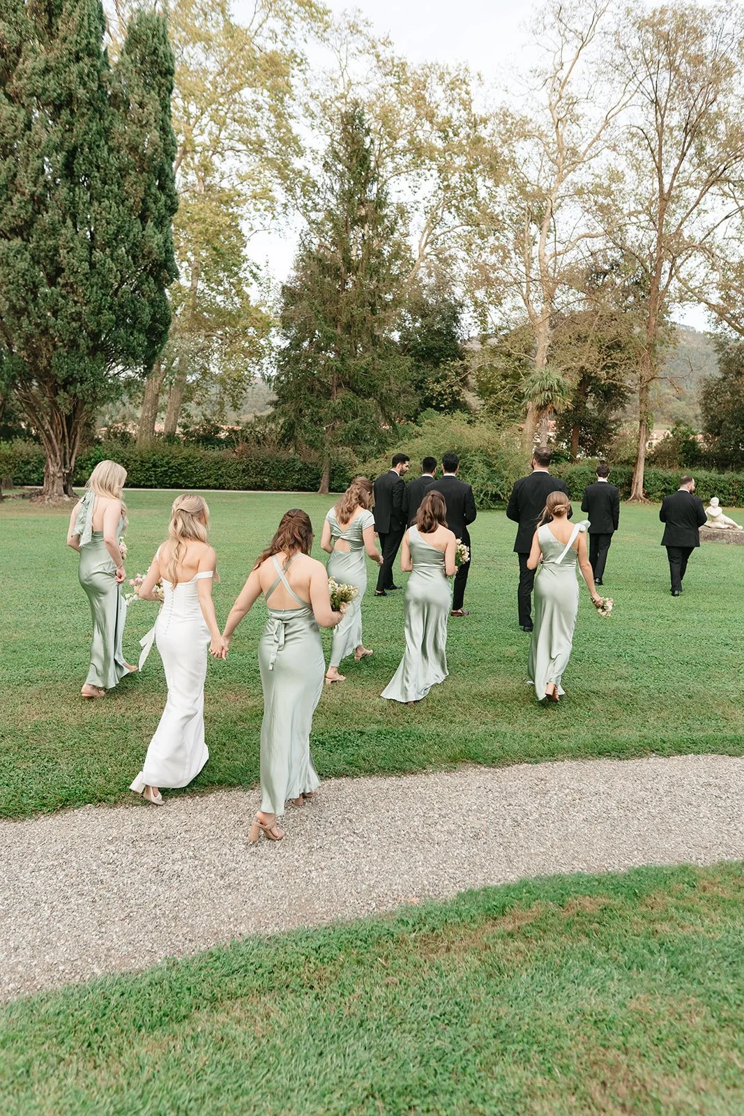 A wedding procession outdoors with bridesmaids in sage green dresses holding bouquets, and groomsmen in black suits with white shirts walking on a grassy area near trees.