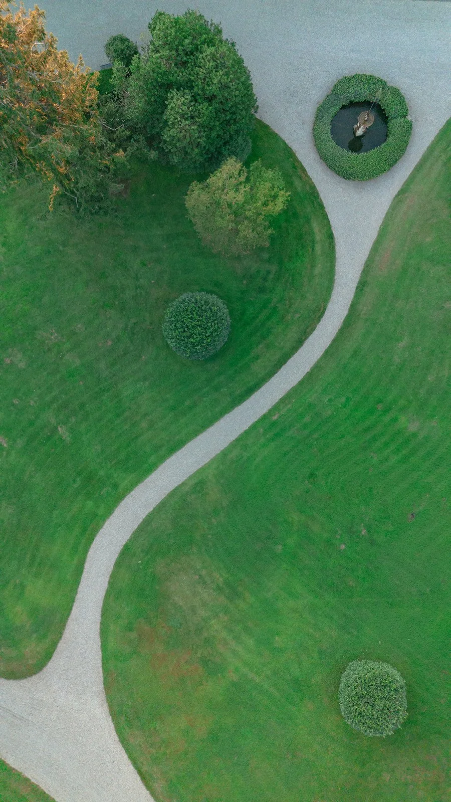 An aerial view of a park with a curved gravel path, green grass, and trees and bushes.