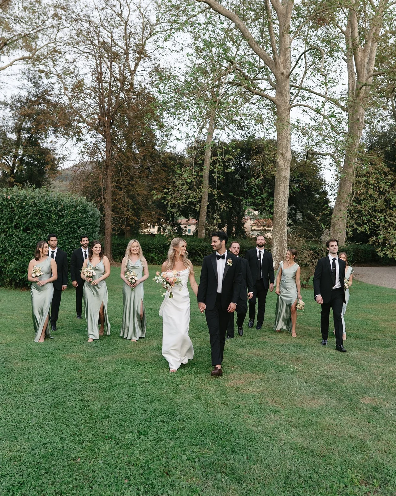 A bride and groom walking hand in hand in a garden with bridesmaids and groomsmen following behind.