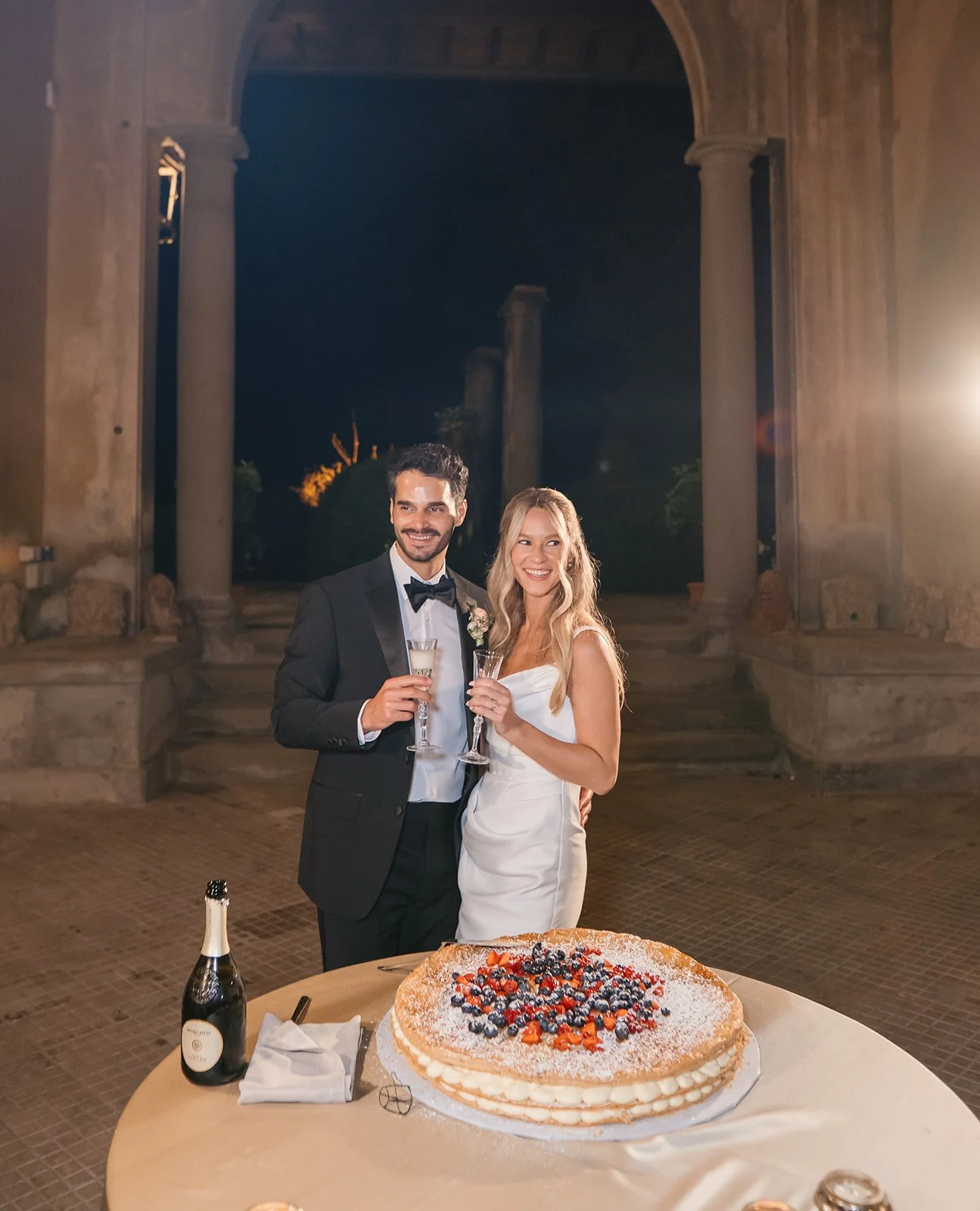 A newlywed couple dressed in wedding attire celebrating at their wedding reception. The groom wears a tuxedo with a bowtie, and the bride wears a white wedding dress. They are holding champagne glasses and standing behind a table with a large, decora