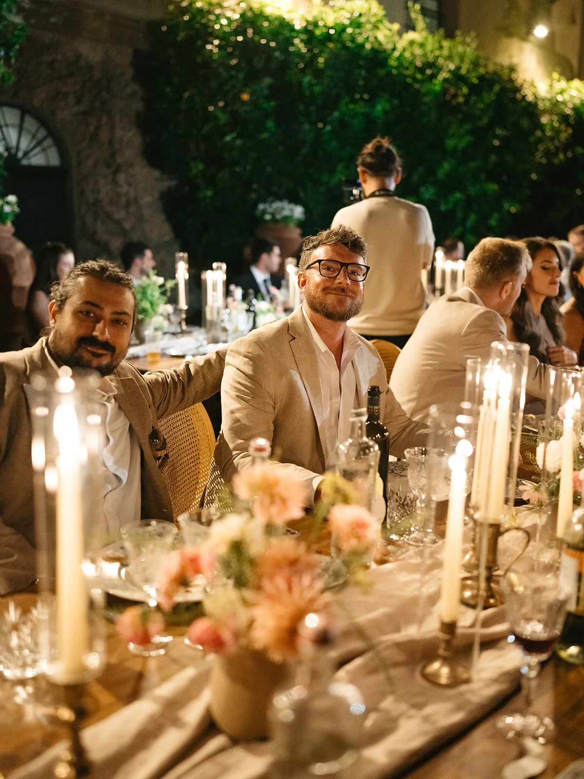 People sitting at a decorated dinner table with candles and flowers, attending an outdoor evening event or celebration.