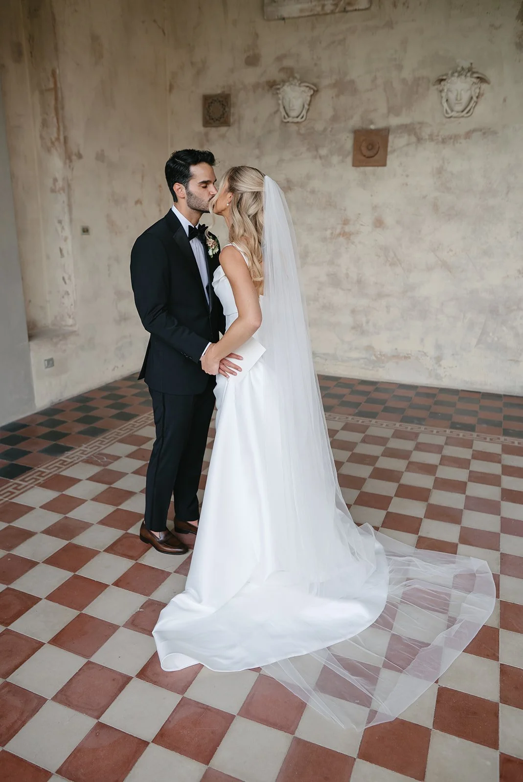A bride and groom kissing indoors on a checkered tile floor, with a textured wall and decorative wall sculptures in the background.