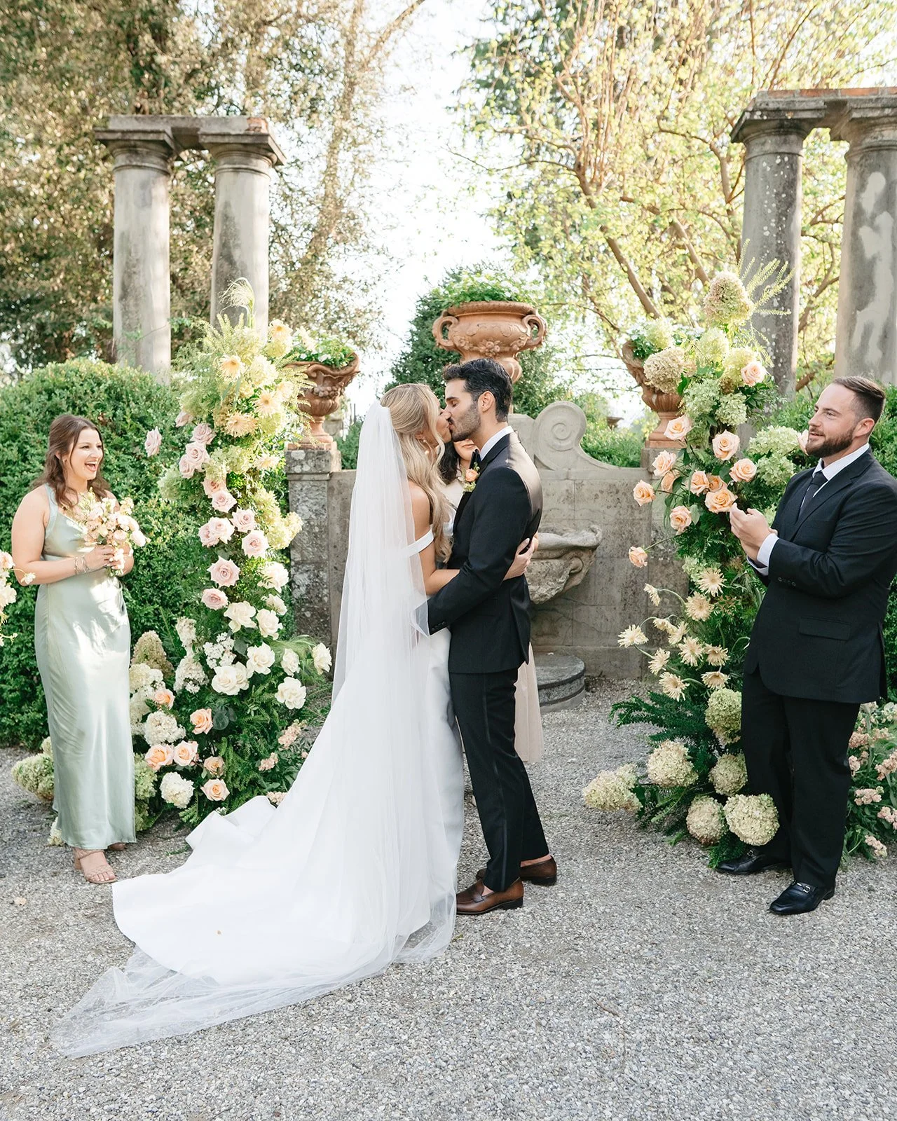 A bride and groom kiss during their outdoor wedding ceremony, surrounded by floral arrangements and greenery, with a bridesmaid smiling and clapping, and a groomsman taking a photo.