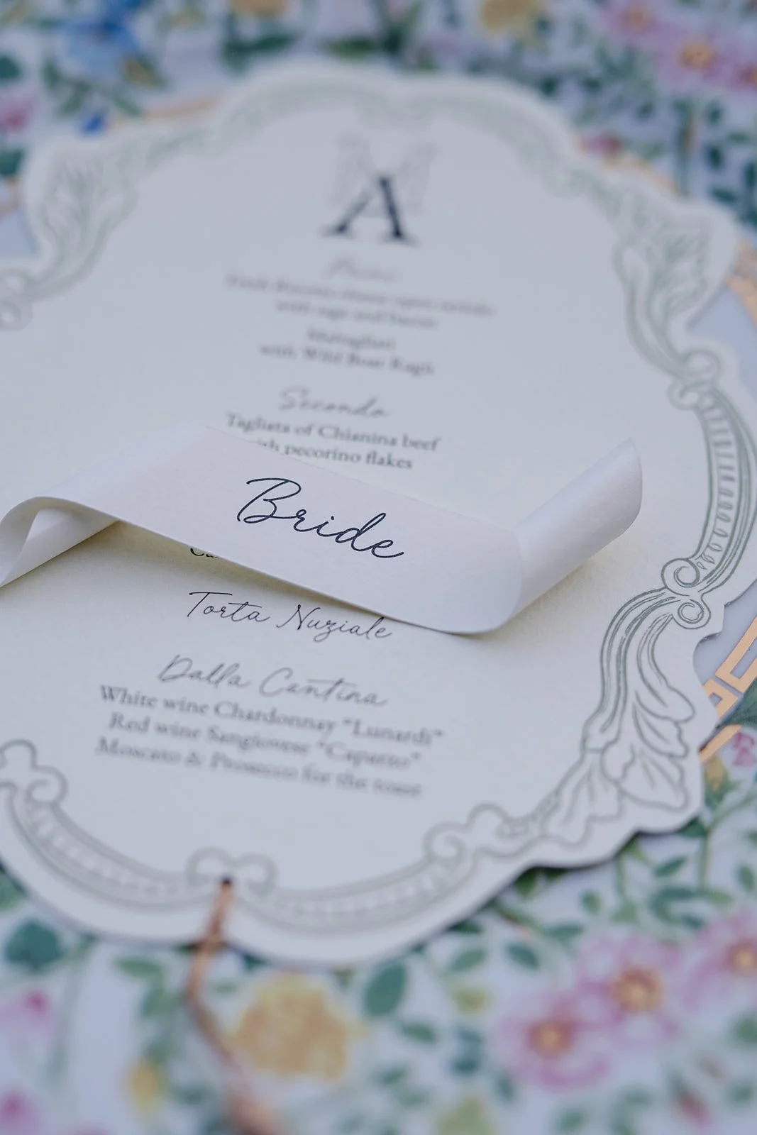 Close-up of a wedding menu with a scroll-like paper, marked 'Bride', resting on floral patterned fabric. The menu lists starter, main course, and wine options.