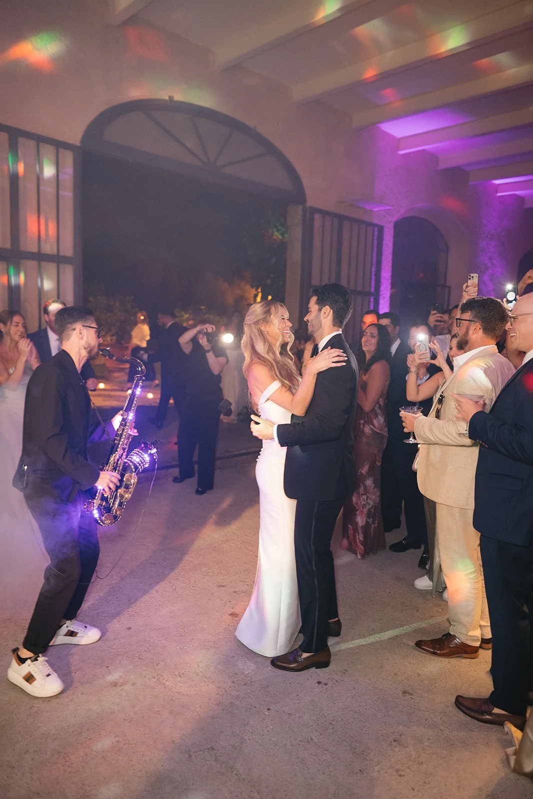 A bride and groom dancing at their wedding reception with guests watching and laughing, while a saxophonist performs nearby, in a decorated indoor venue with colorful lighting.
