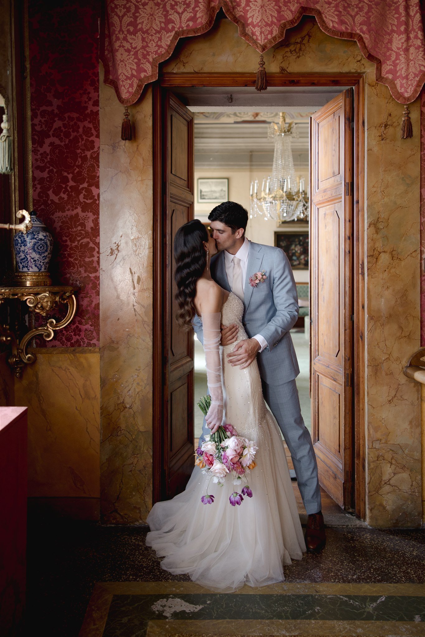A bride and groom sharing a kiss, standing in an arched stone doorway with greenery and a fountain in the background, during a wedding ceremony.