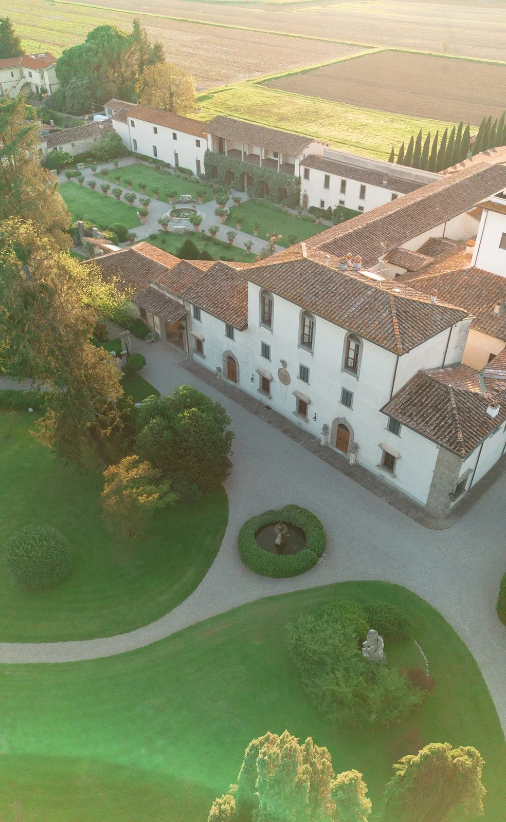 An aerial view of a historic white building with a red-tiled roof, surrounded by a manicured garden with bushes, trees, and a small fountain, located in a rural area with fields in the background.
