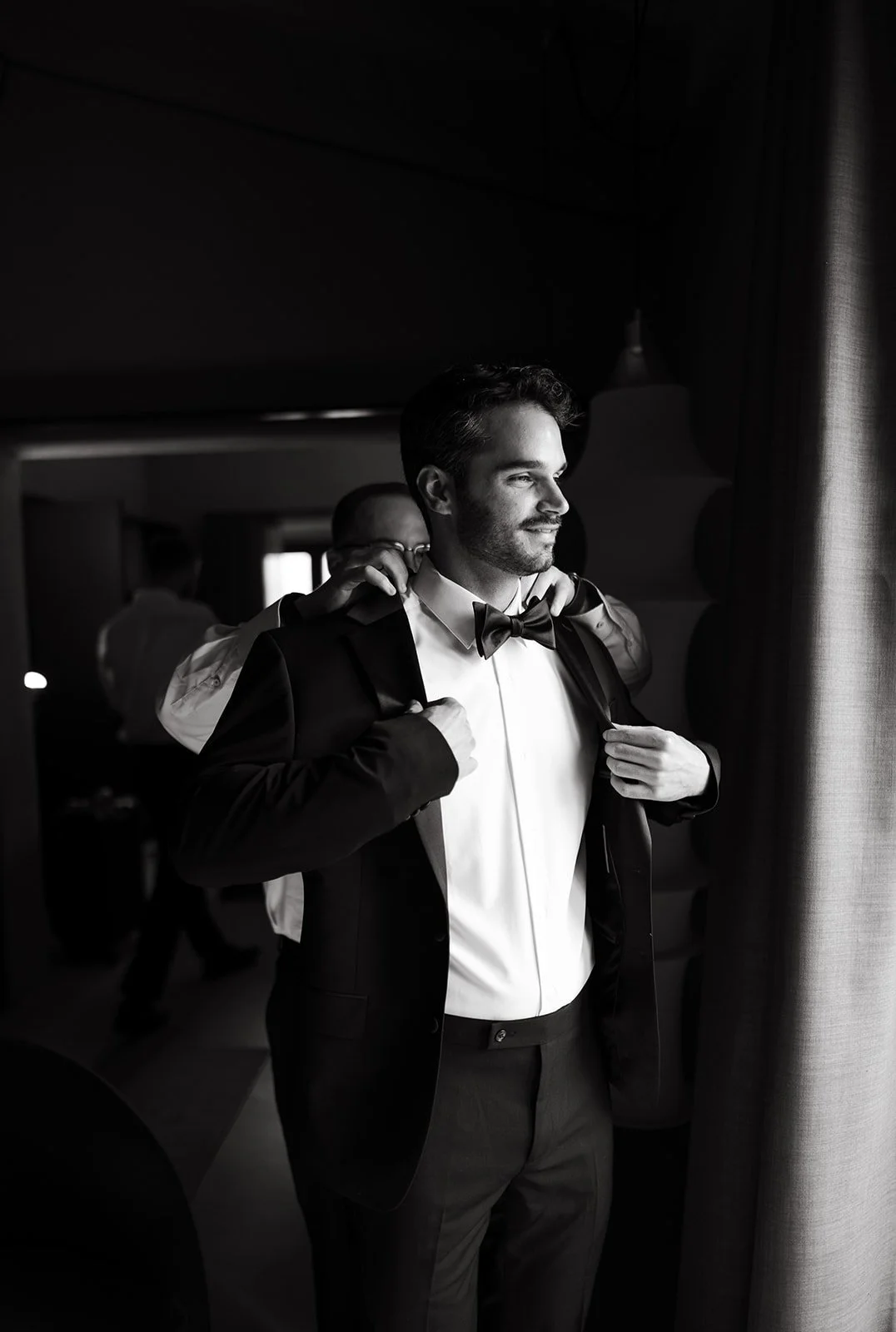 A man in a tuxedo with a bow tie stands near a window, adjusting his jacket. Another person helps him with his jacket in the background.