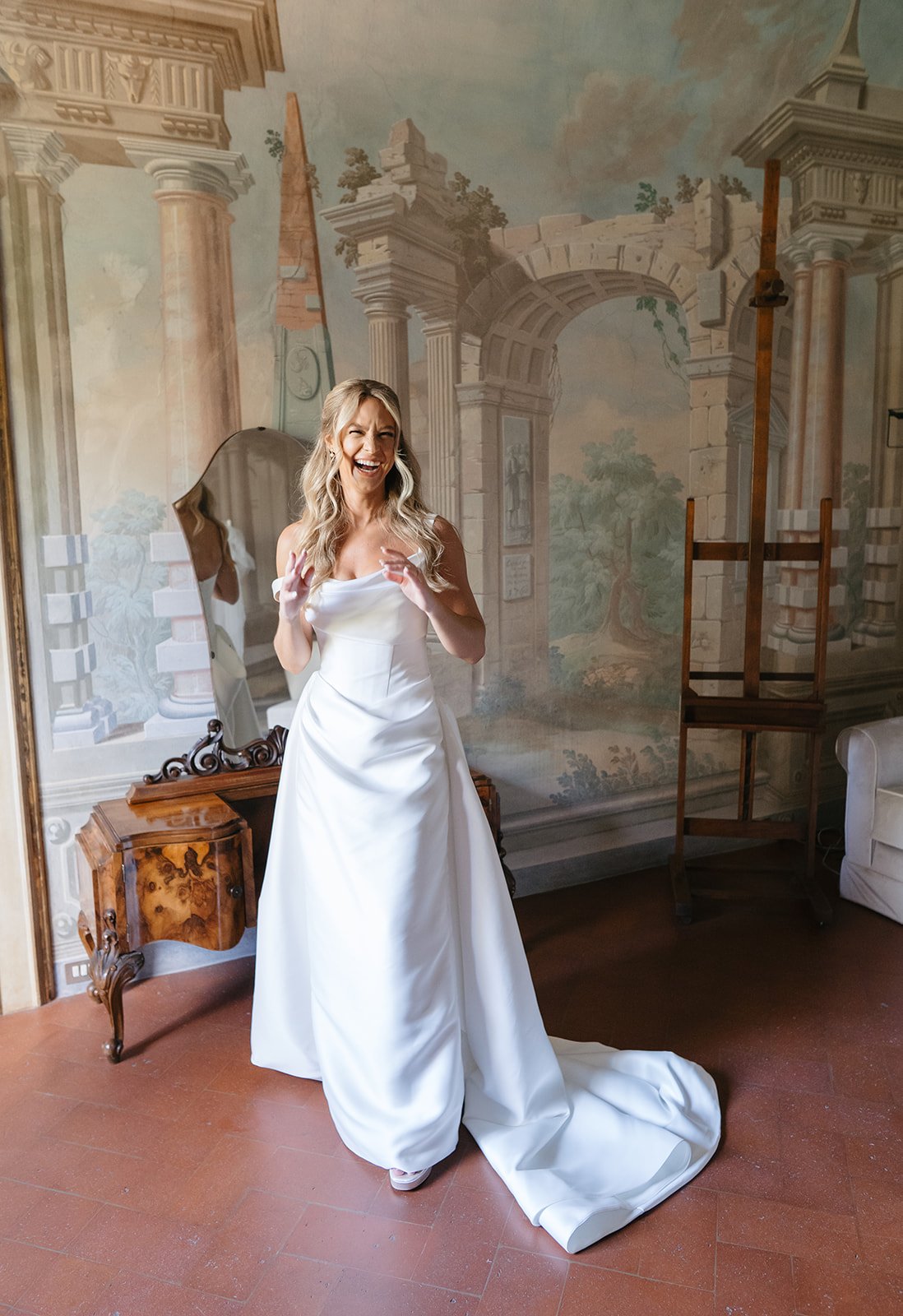 A woman in a white wedding dress standing in front of a painted mural of ancient ruins, smiling and gesturing with her hands.