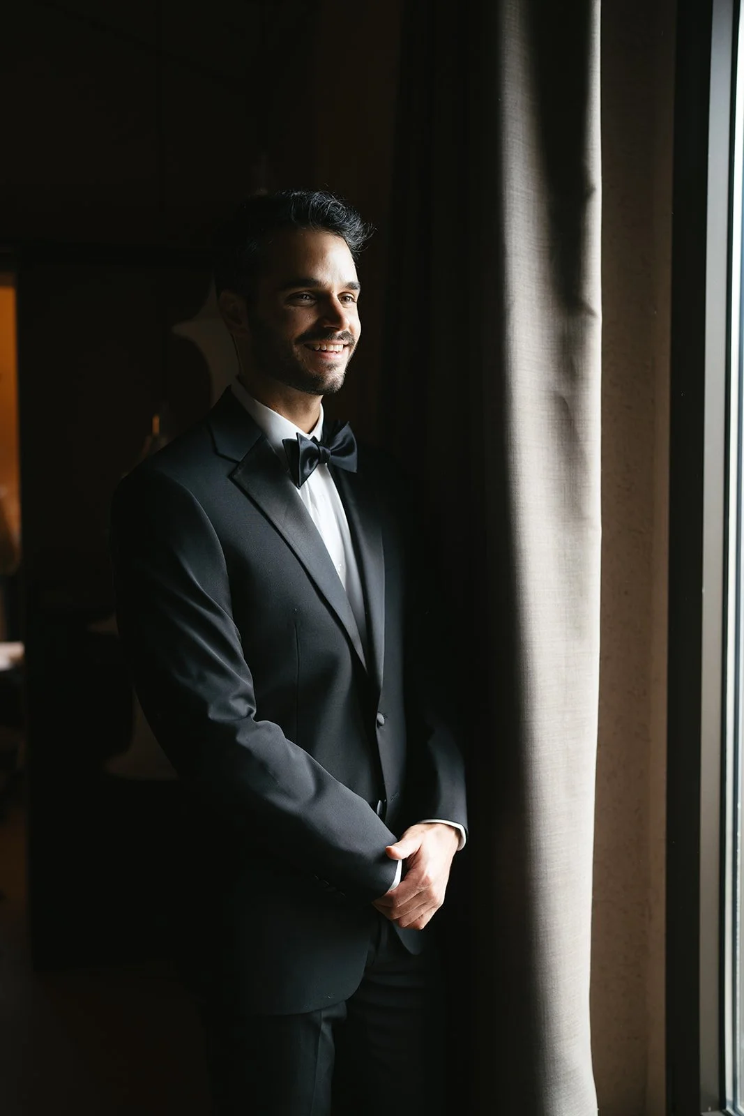 A man in a black tuxedo with a bow tie, smiling and standing by a window with curtains.