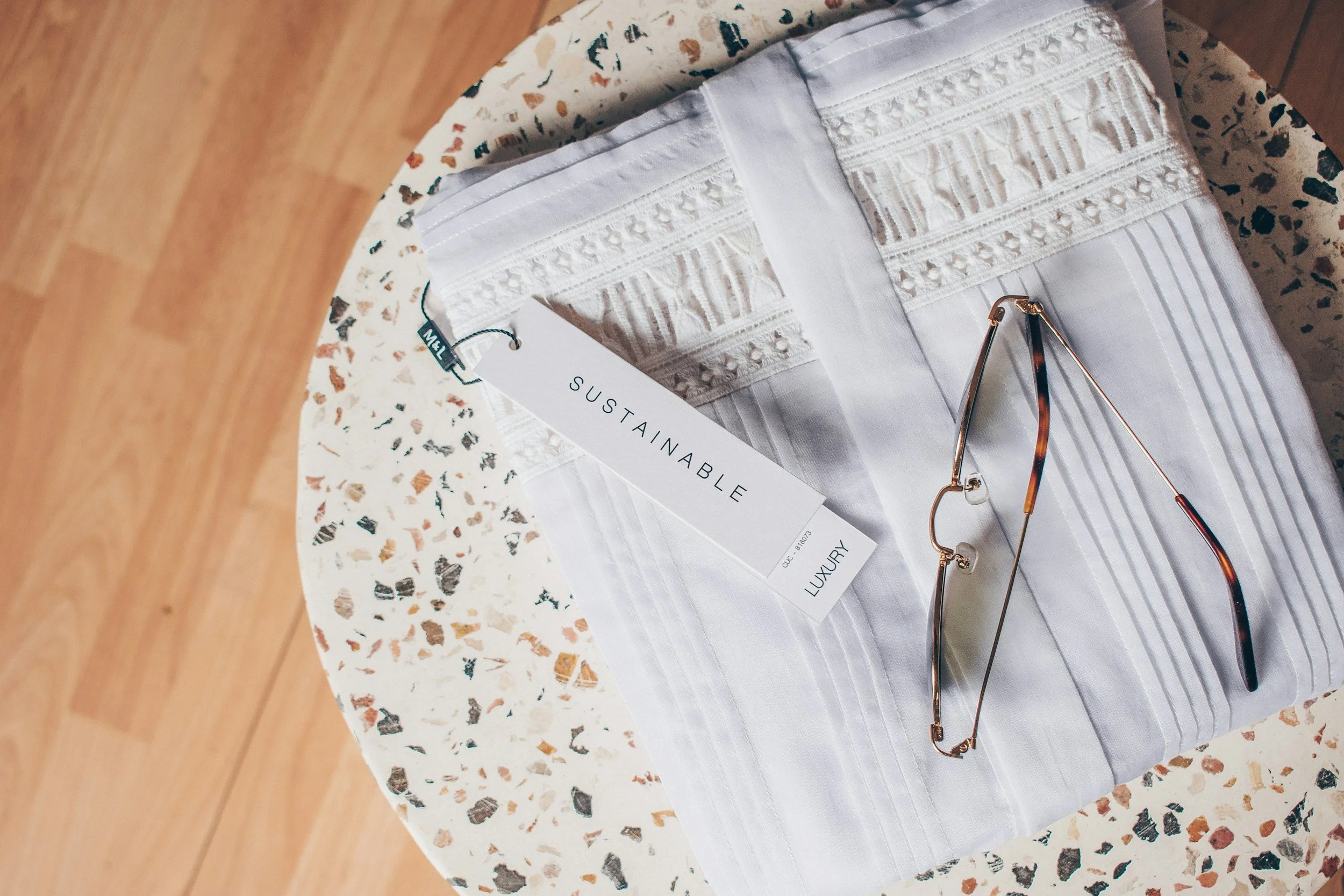 A pair of white fabric pants with intricate lace and embroidery details, a size tag labeled "sustainable luxury," and a pair of tortoise shell glasses on a terrazzo table with a wooden floor in the background.