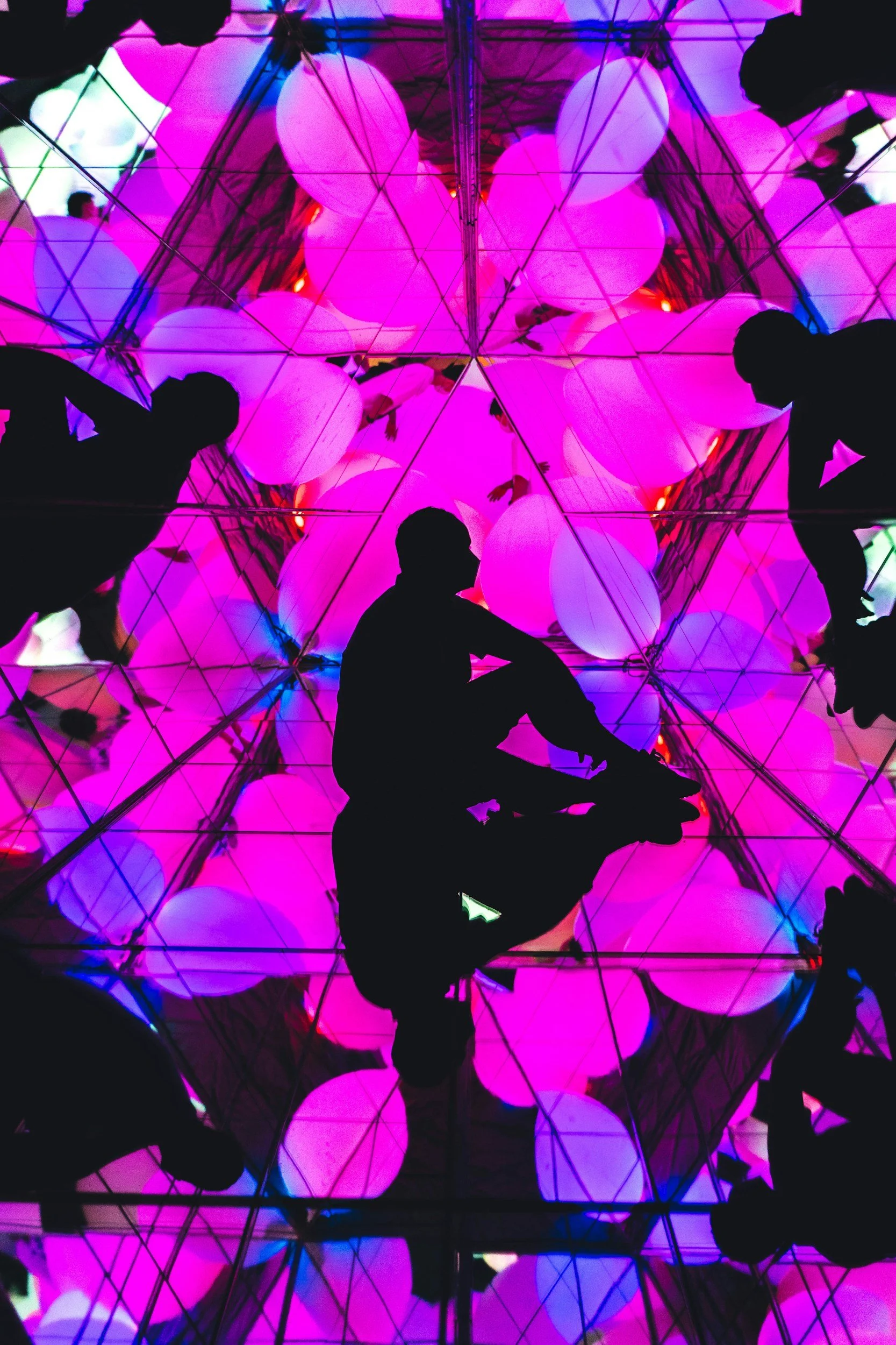 Silhouettes of people sitting and leaning against a reflective surface with colorful, pink, purple, and blue glowing orbs and geometric reflections.