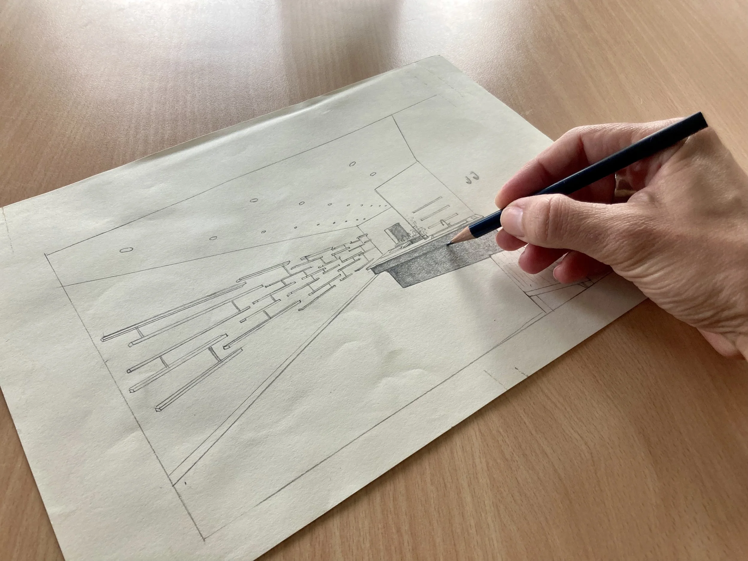 A hand drawing an interior architectural design on paper with a pencil, showing a retail space with bespoke counter and shelving