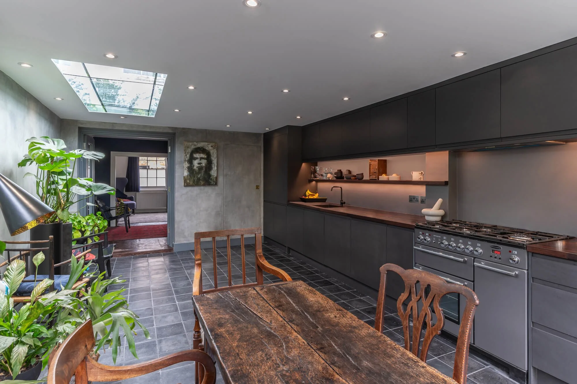 Modern kitchen with black cabinets, wooden countertop, gray tiled floor, and a dining table with wooden chairs. Plants are on the left, and a skylight and recessed lighting illuminate the space.
