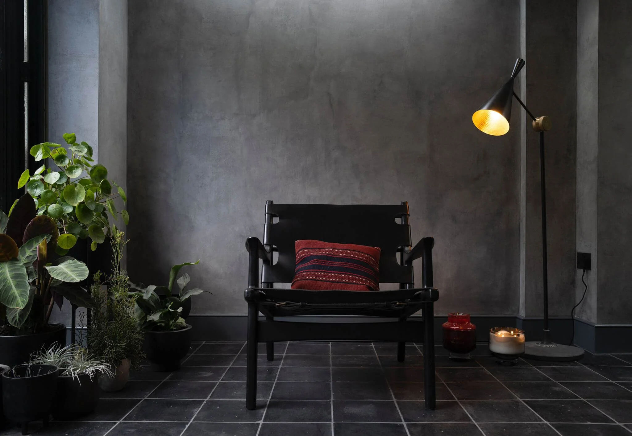 A black bench with a red cushion in front of a gray wall, surrounded by potted green plants on the left, and a floor lamp with a yellow interior on the right, with candles on the floor near the lamp.