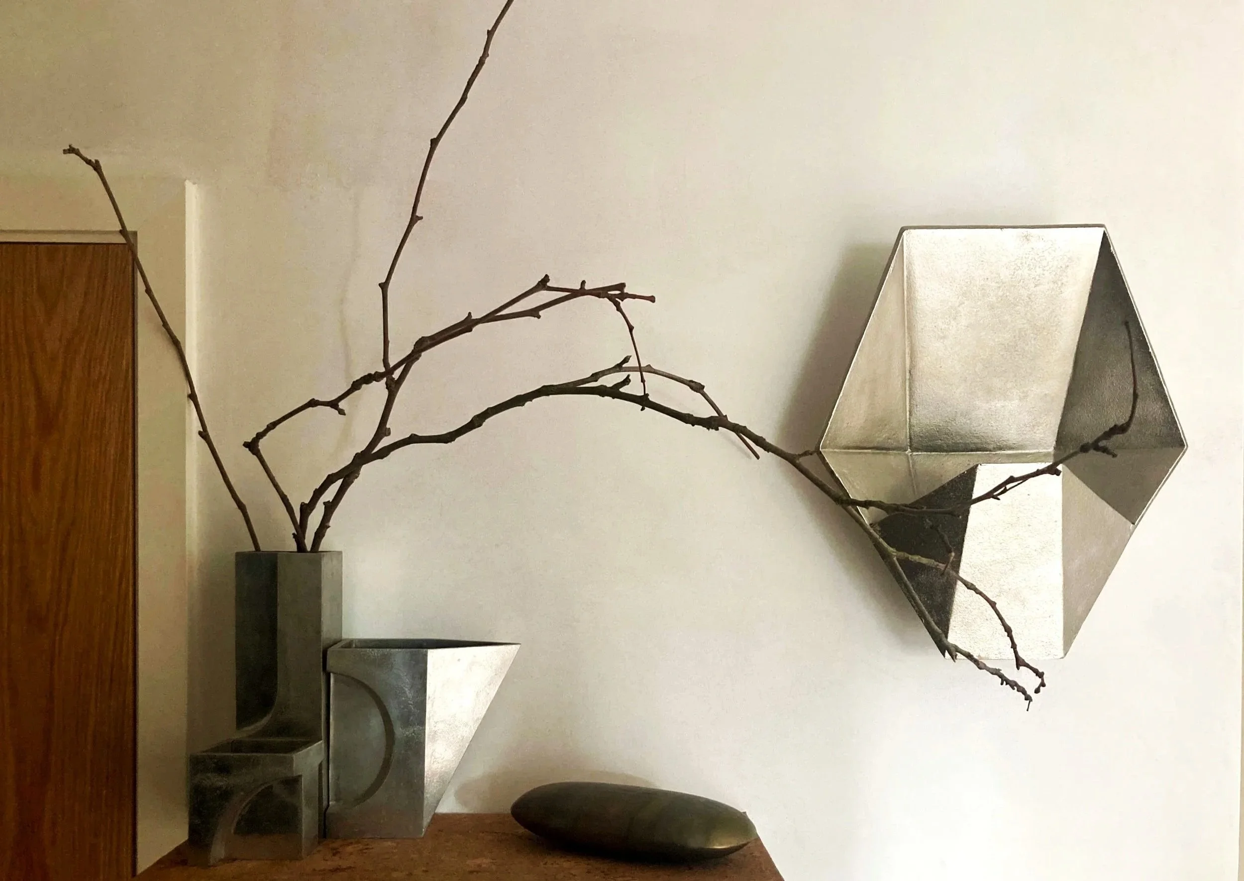 Decorative interior with a vase holding bare branches, geometric wall sconces, and a smooth black stone on a wooden surface.