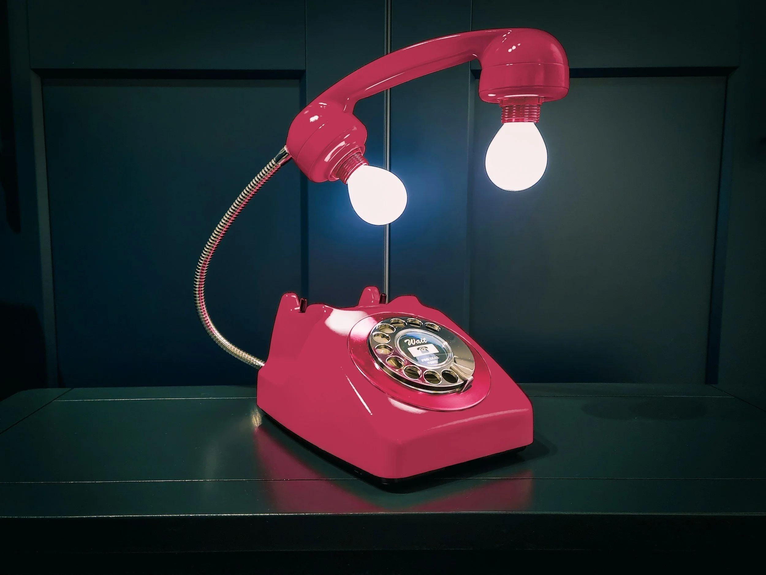 Pink rotary phone with a lamp shaped like its handset, standing on a black surface against a dark blue background.